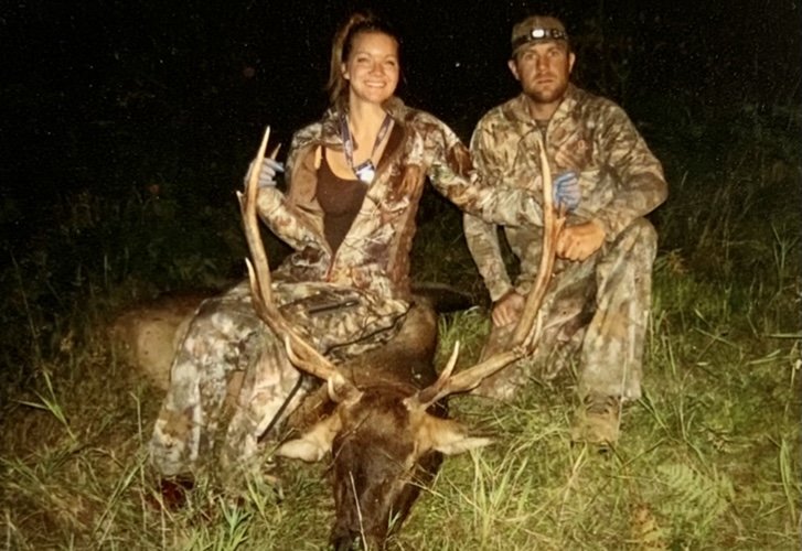 Two hunters in camouflage gear kneeling beside a large elk with impressive antlers on a grassy field at night.