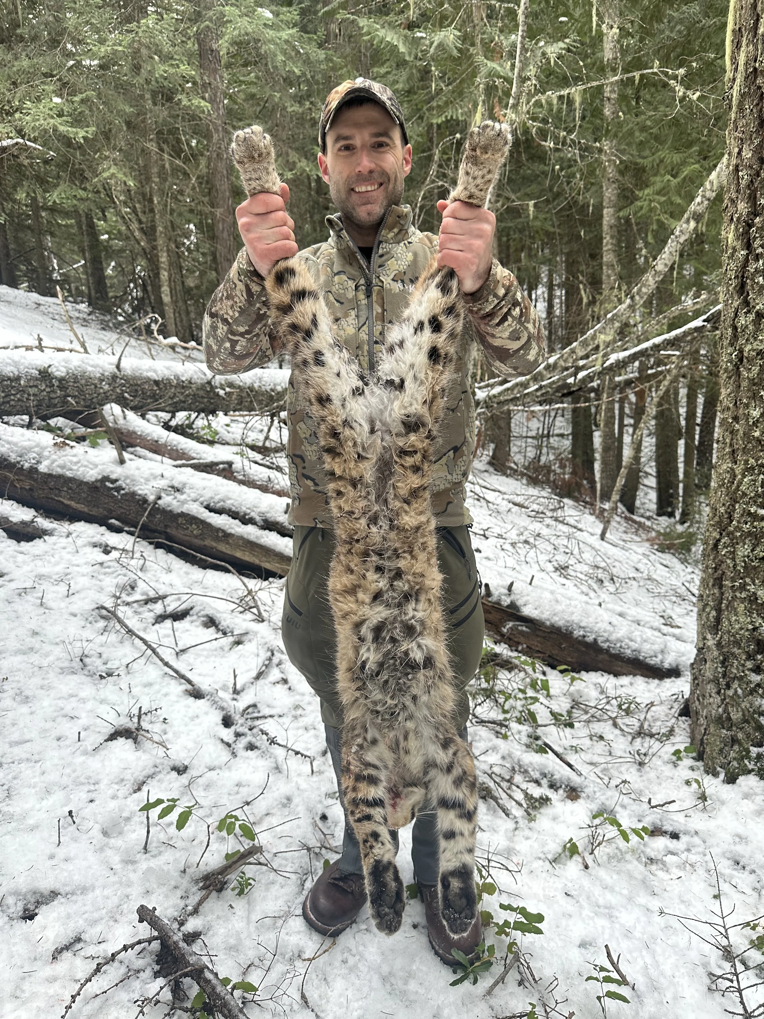 A man dressed in camouflage holding a large snow leopard pelt by its front legs in a snowy forest.