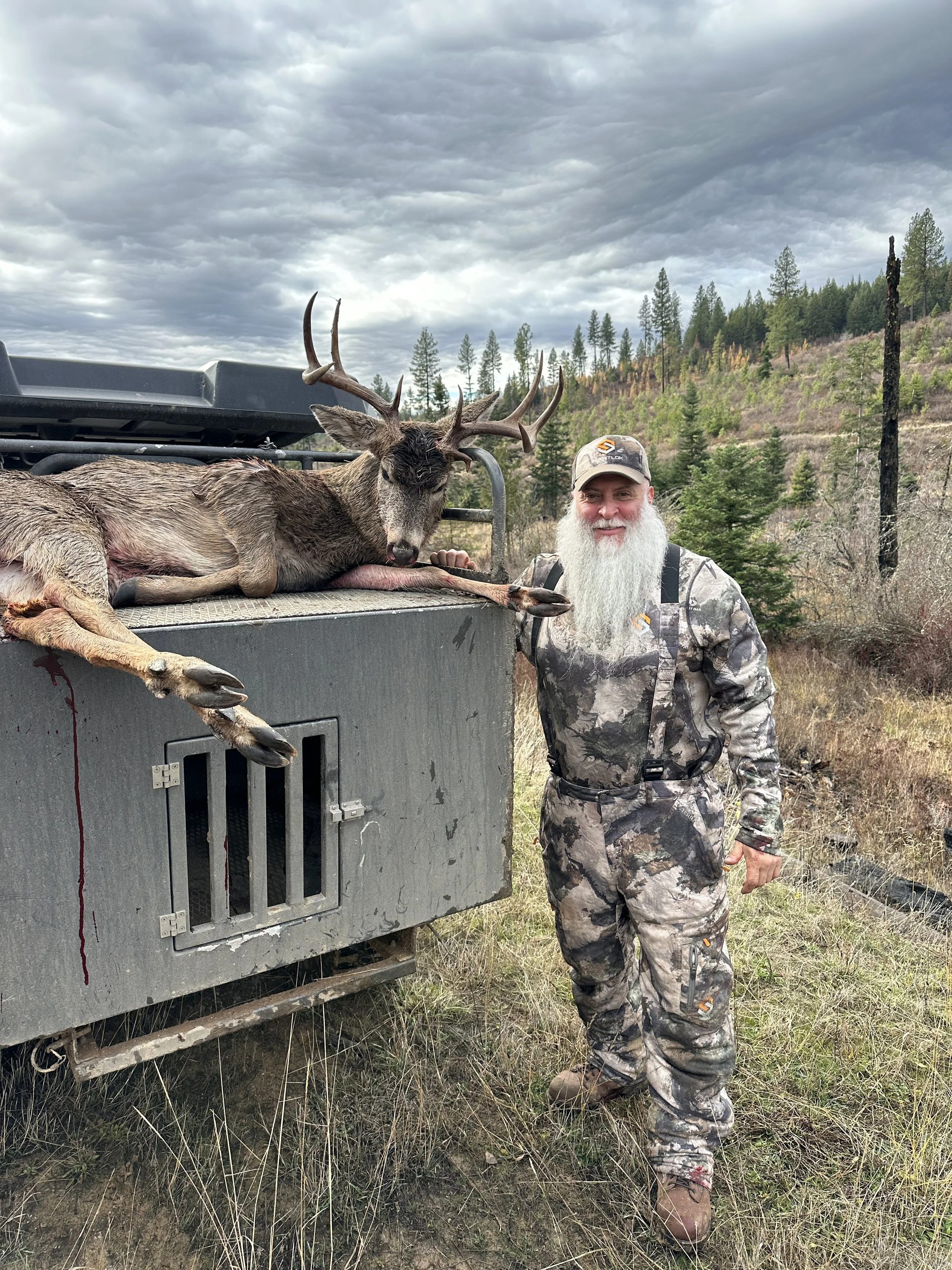 A man dressed in camouflage clothing stands outdoors next to a large truck, smiling, with a freshly hunted deer with antlers lying on top of the truck.