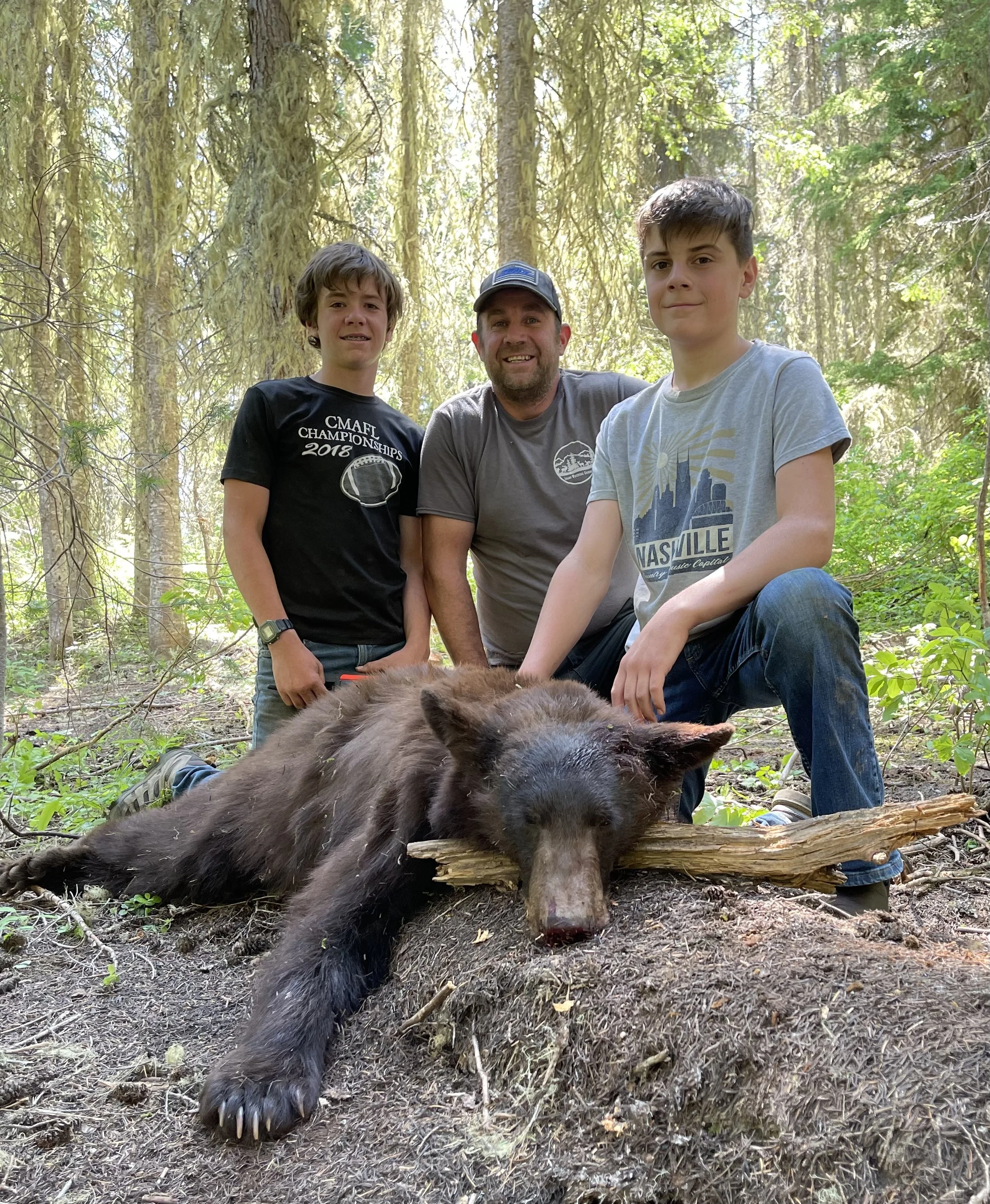 Three people, two boys and an adult man, kneeling beside a dead wild boar in a forest, with the boys touching its head, while the man is smiling, surrounded by trees and foliage.