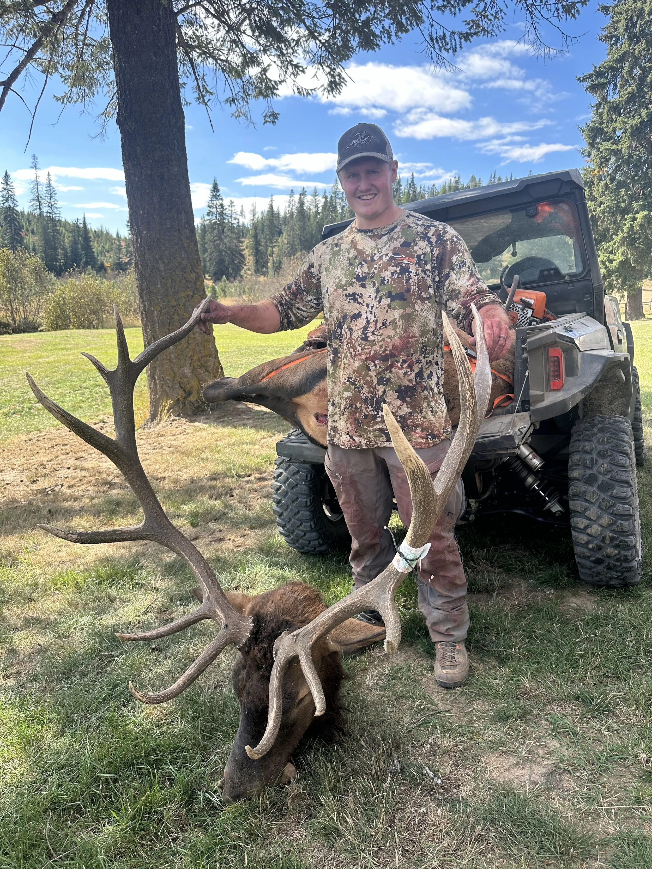 A man in camouflage clothing standing outdoors on grass, holding the antlers of a large elk with a dead elk's head on the ground. There is a utility vehicle behind him and trees in the background under a partly cloudy sky.