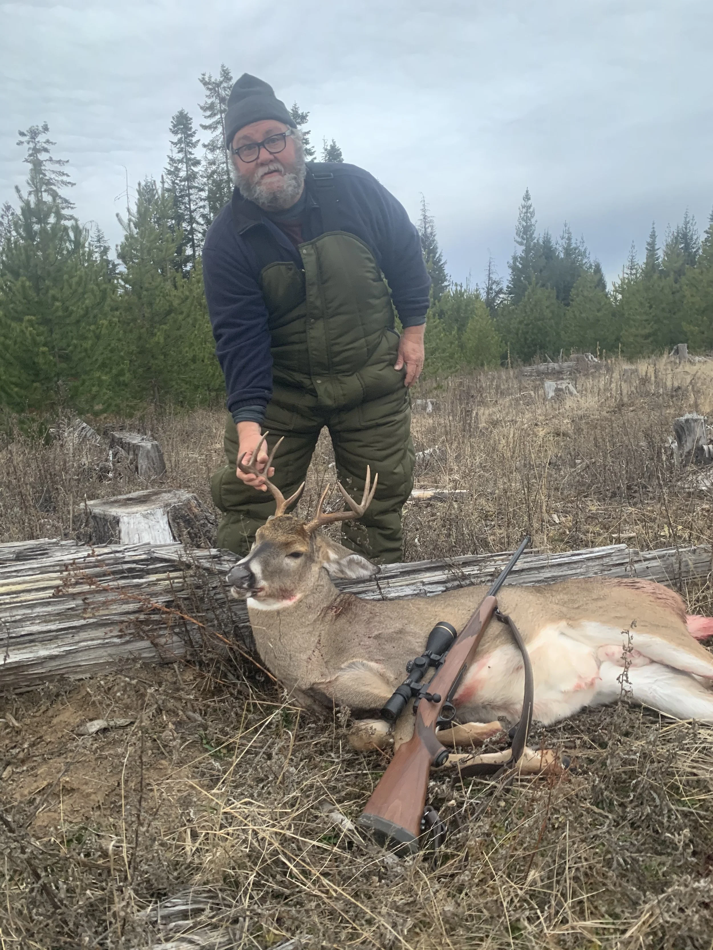 Man with glasses and beard in camouflage outdoor clothing posing next to a buck with antlers. The buck is lying on the ground with a rifle and scope resting on it. Forested area with trees and logs in the background.