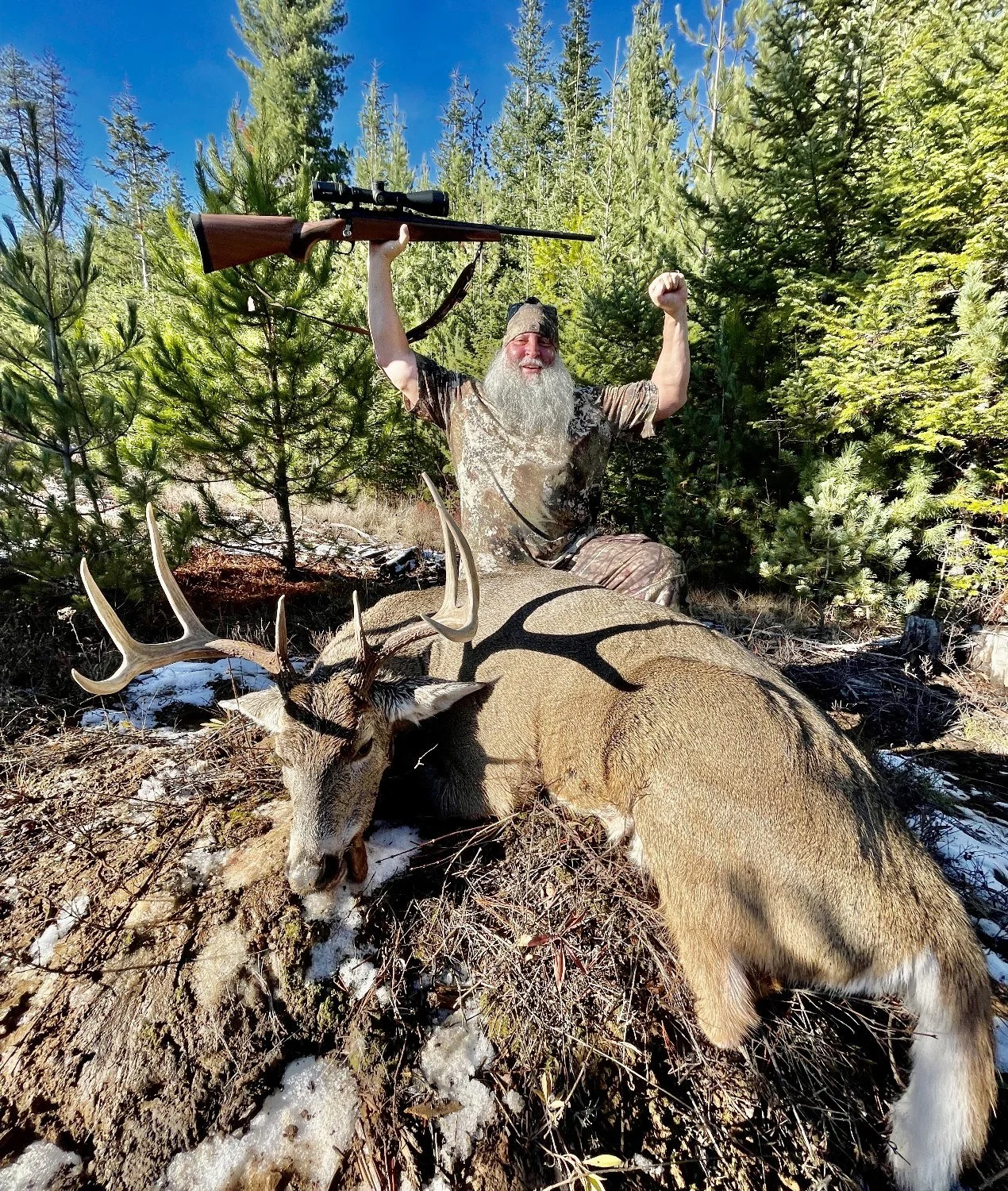A man with a long gray beard celebrating after a successful hunt in a forest, holding a rifle aloft while sitting behind a large deer with antlers, surrounded by green trees under a clear blue sky.