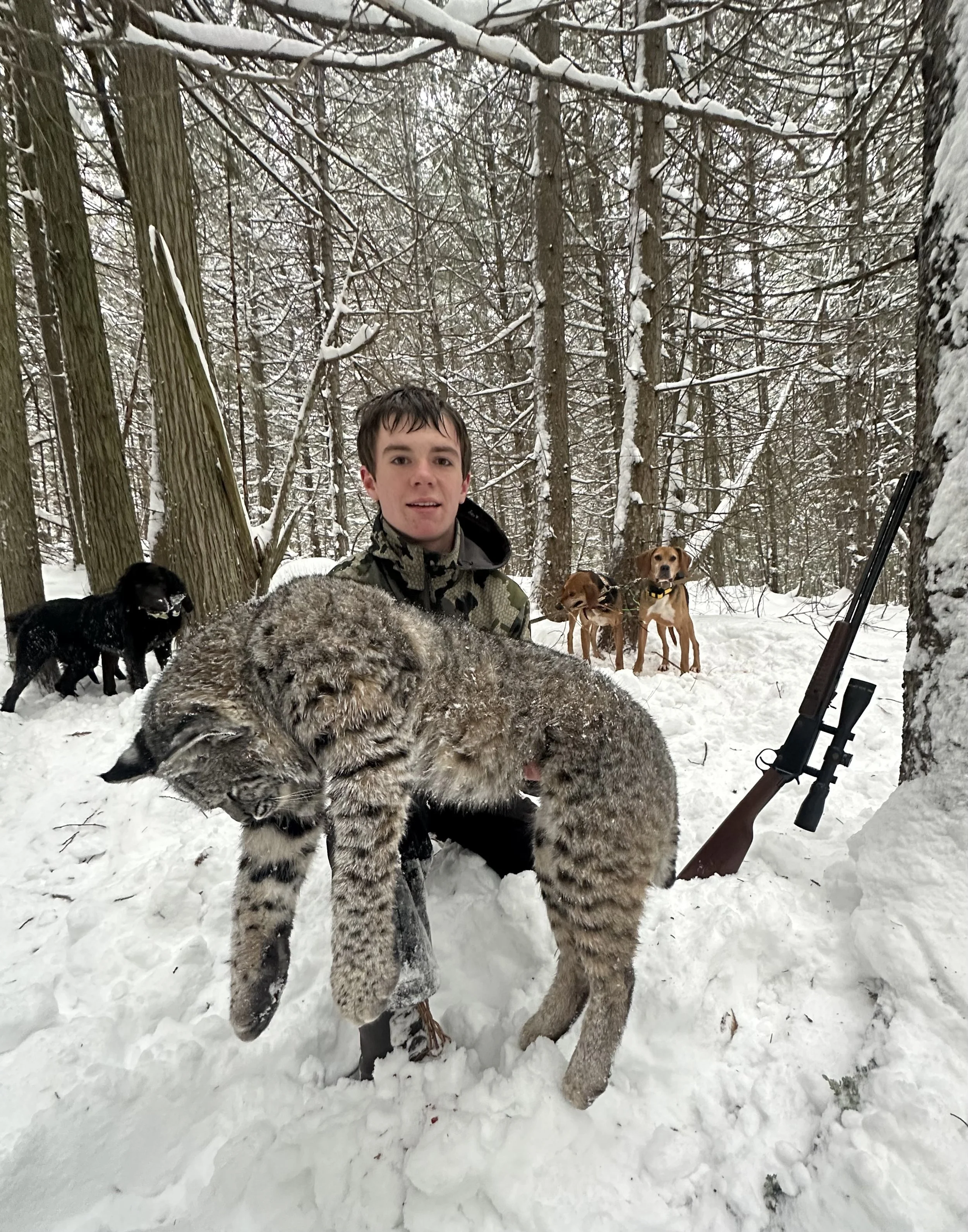 A young man in a camo jacket standing in a snowy forest, holding a snow-covered snow leopard. Three dogs and a rifle are also visible in the background.