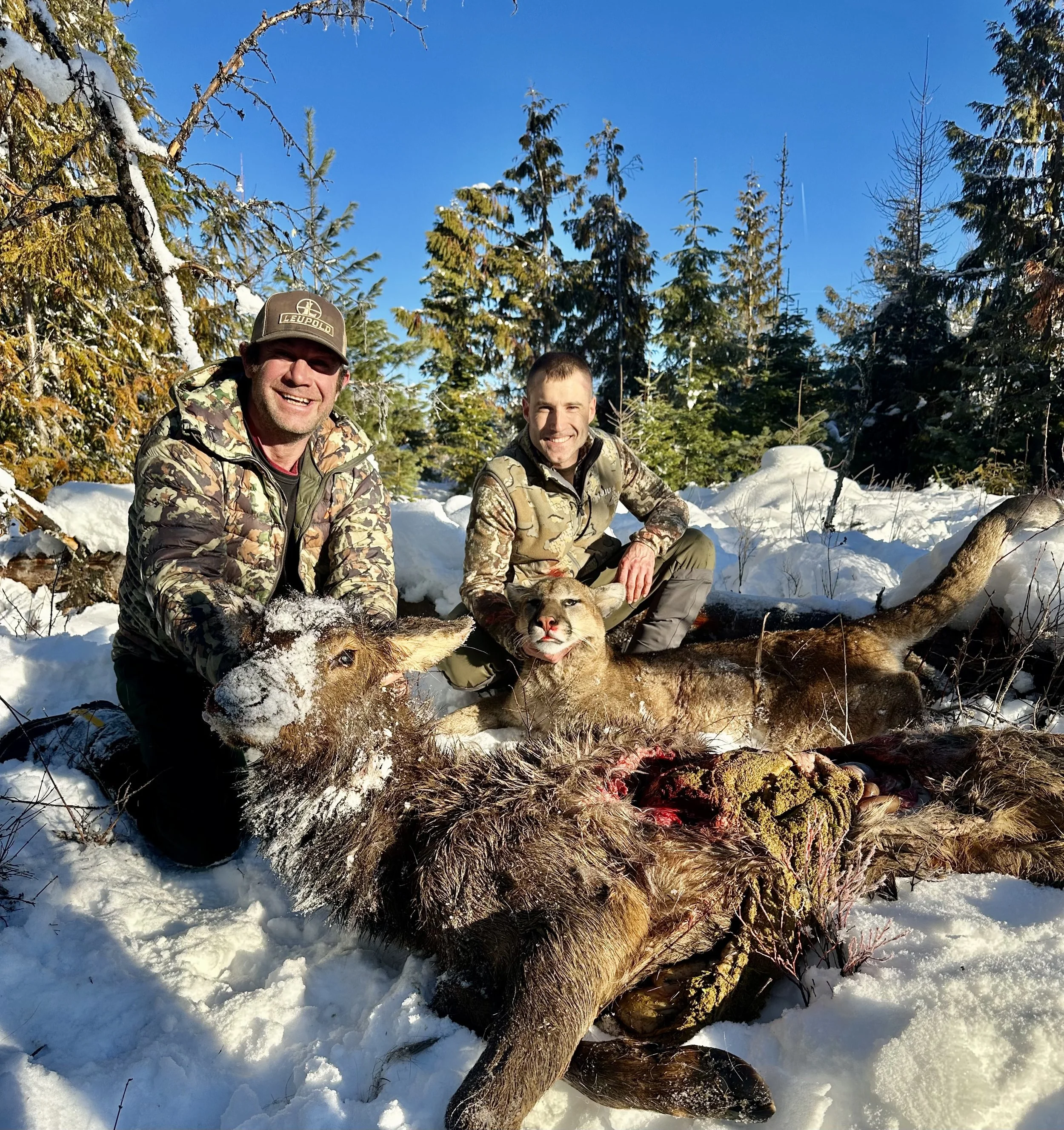 Two men in camouflage clothing with a dog and a dead elk in a snowy forest landscape on a sunny day.