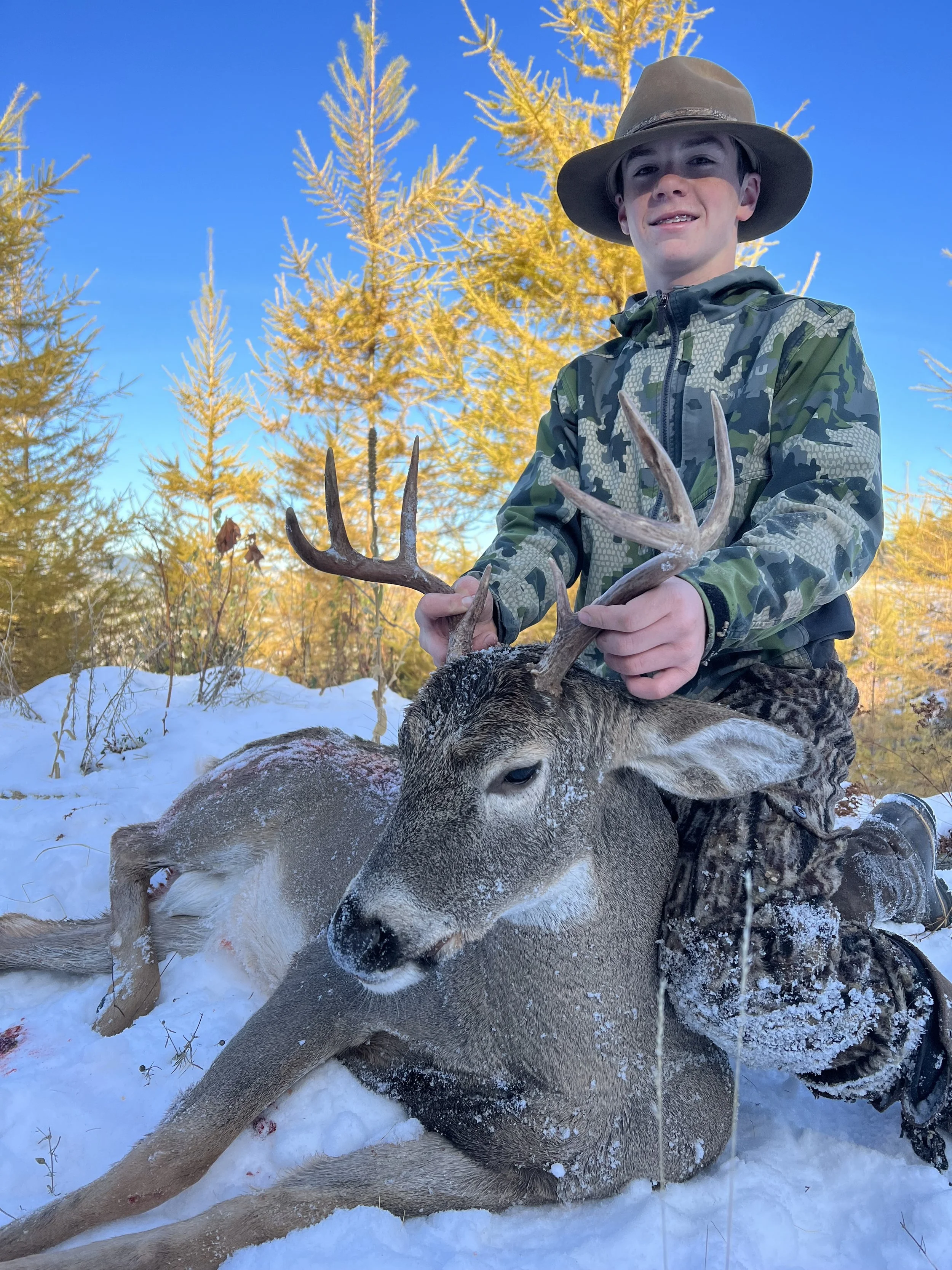 A boy in camouflage and a wide-brimmed hat kneeling in snow, holding antlers of a freshly hunted deer with a snowy forest background.