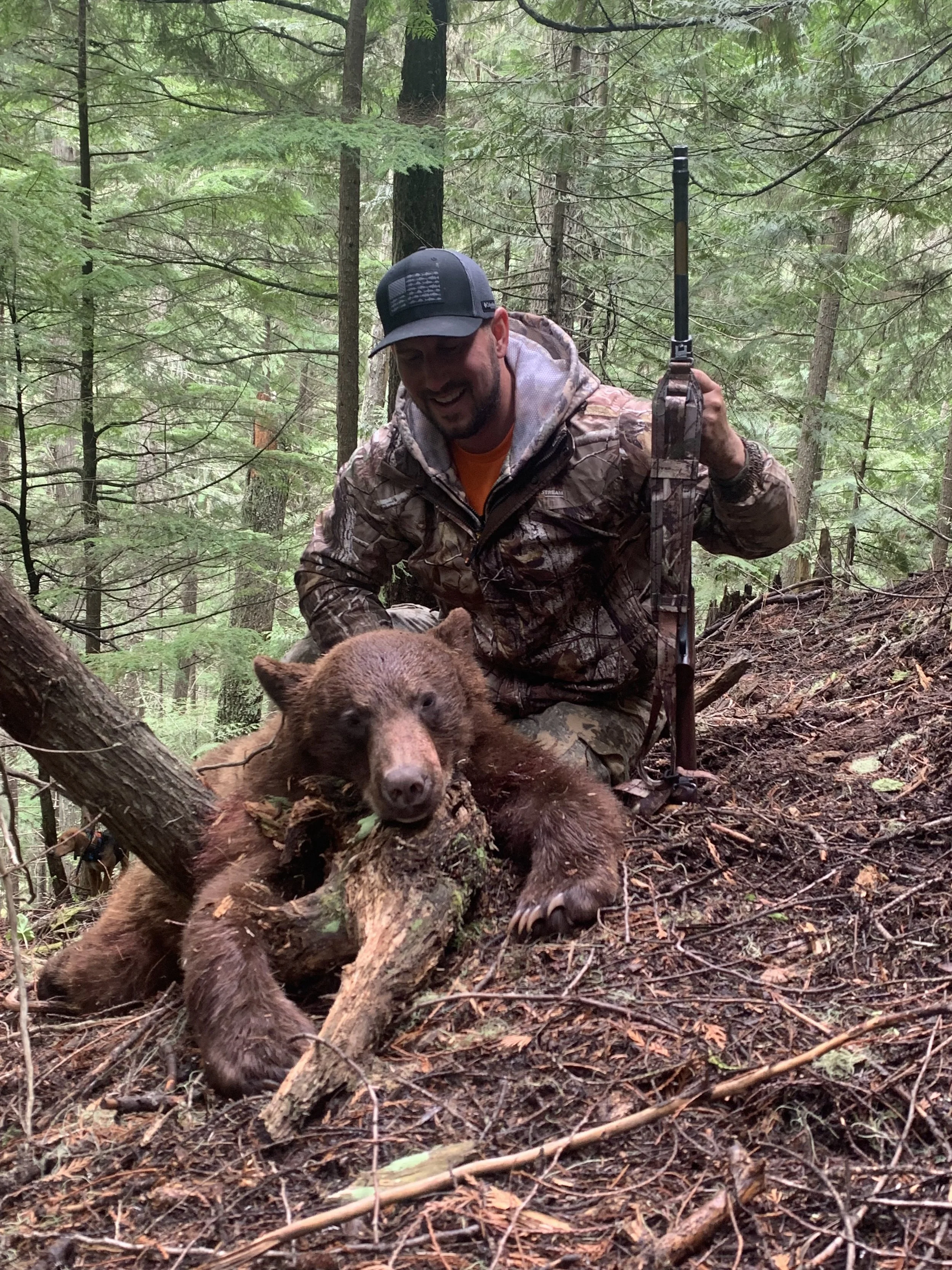 A man in camouflage clothing kneels in a forest, smiling and holding a rifle, next to a recently hunted brown bear that is lying on the ground with its head resting on a fallen tree.