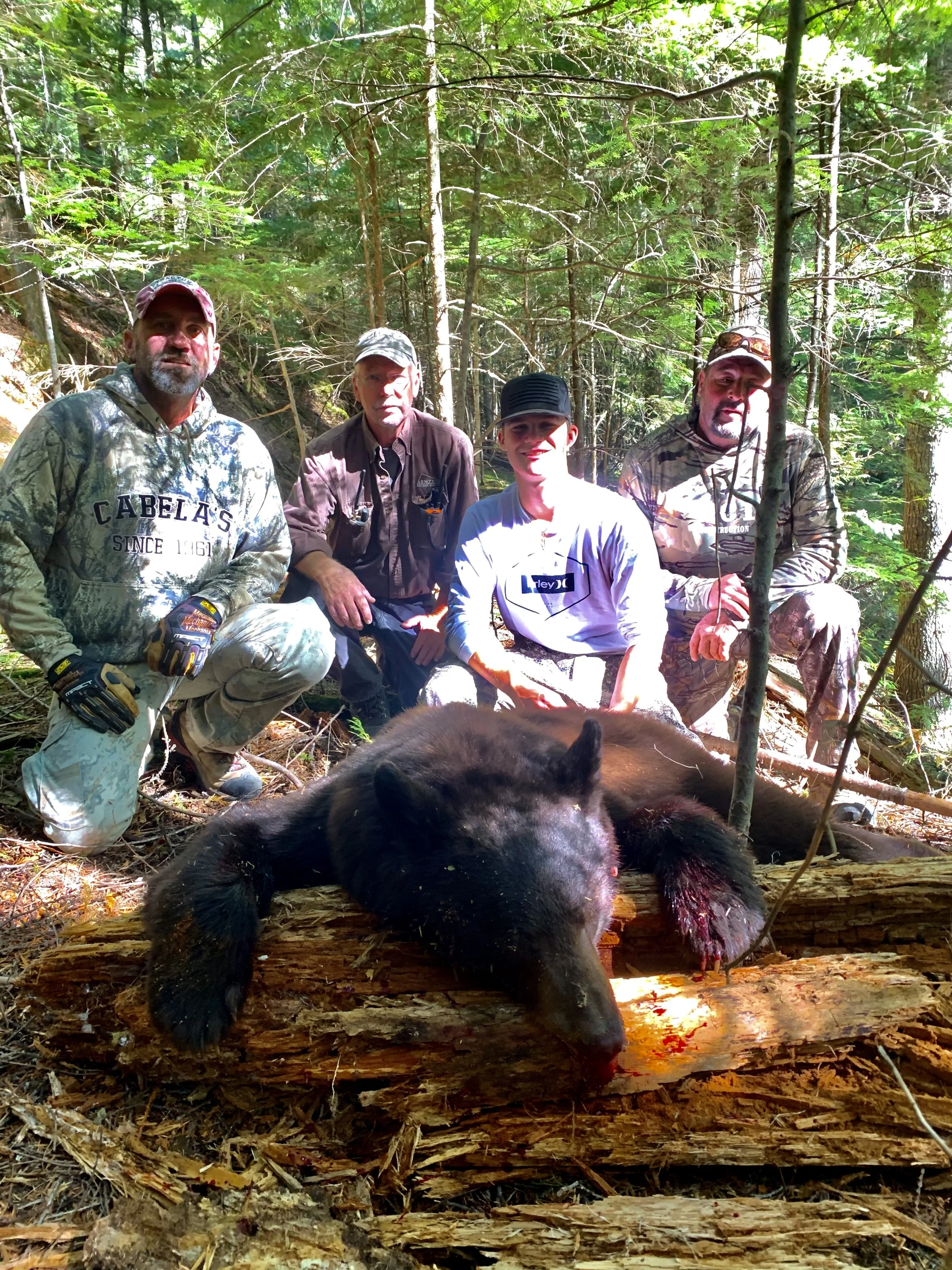 Four hunters in camouflage clothing kneel in a forest behind a large, dead black bear lying on a fallen log.