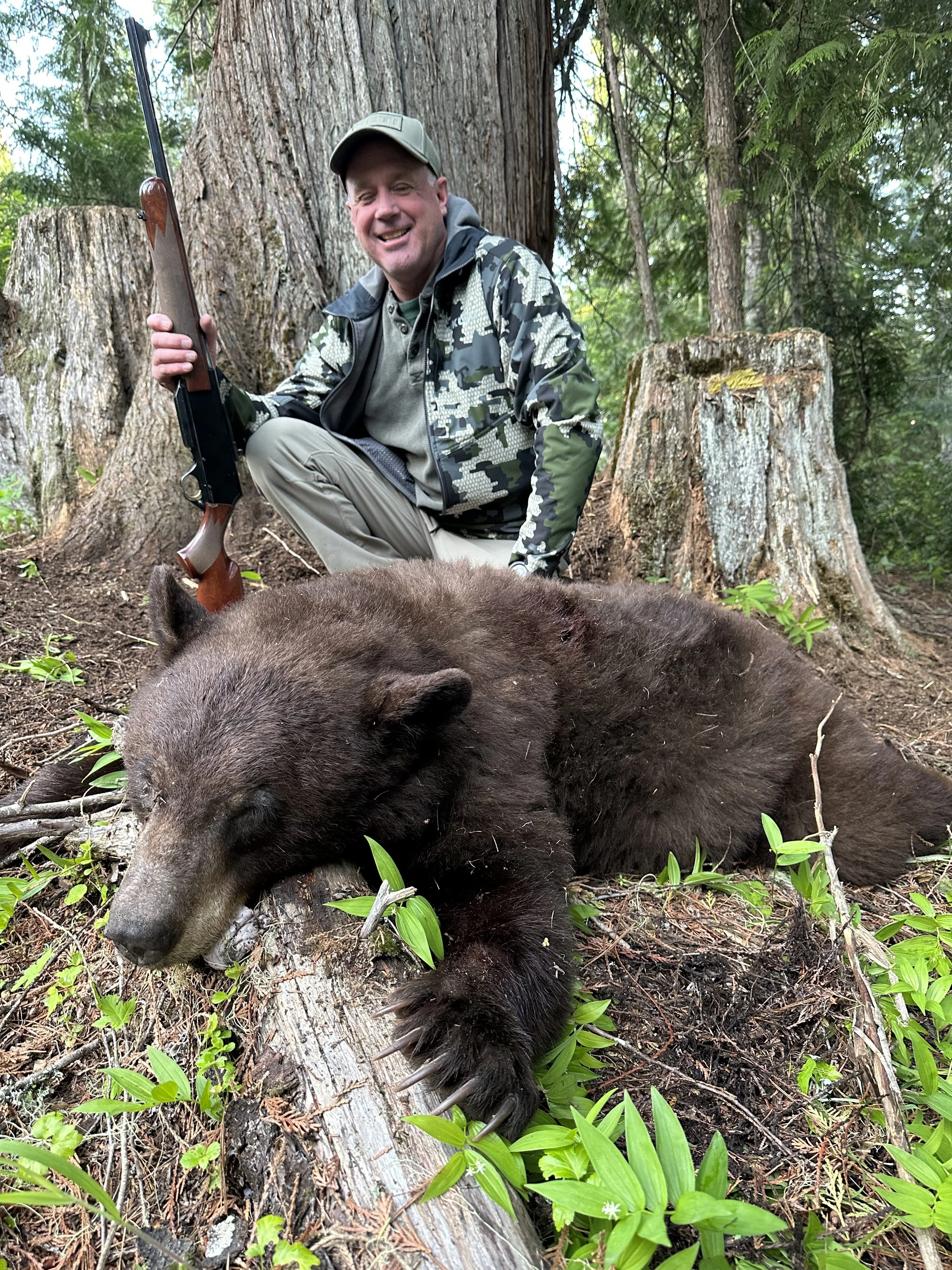 A man in camouflage hunting clothing kneeling next to a large, dead bear in a forested area, holding a rifle, with trees and green foliage around.