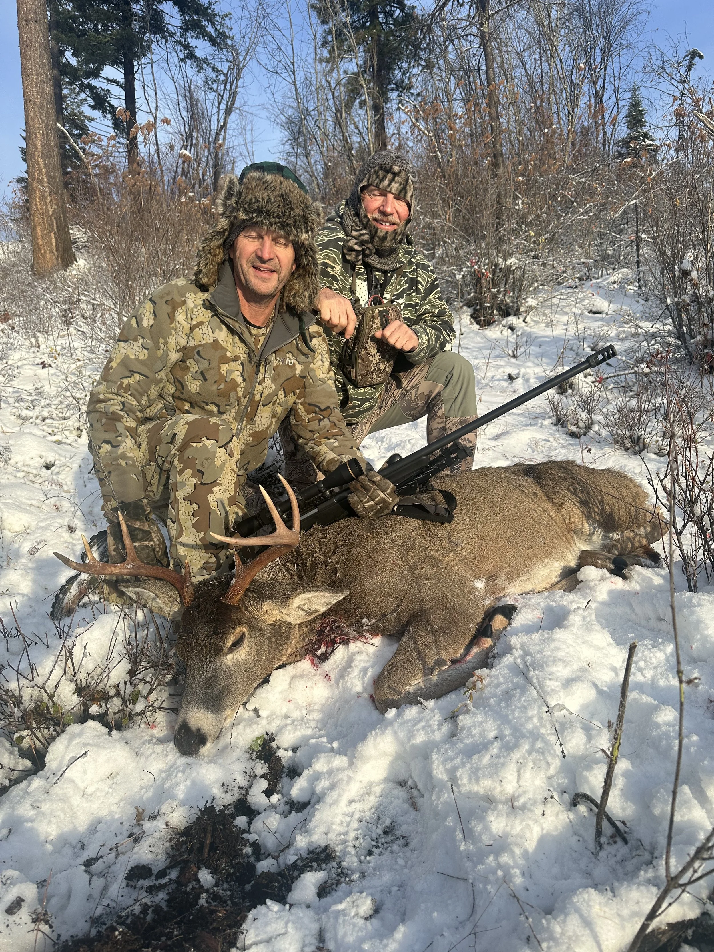 Two hunters in camouflage clothing kneel next to a freshly killed deer in a snowy forest. One man holds a rifle and wears a fur-lined hat, while the other has a camouflage beanie. They appear to be proud of their hunt, smiling and posing with the dee