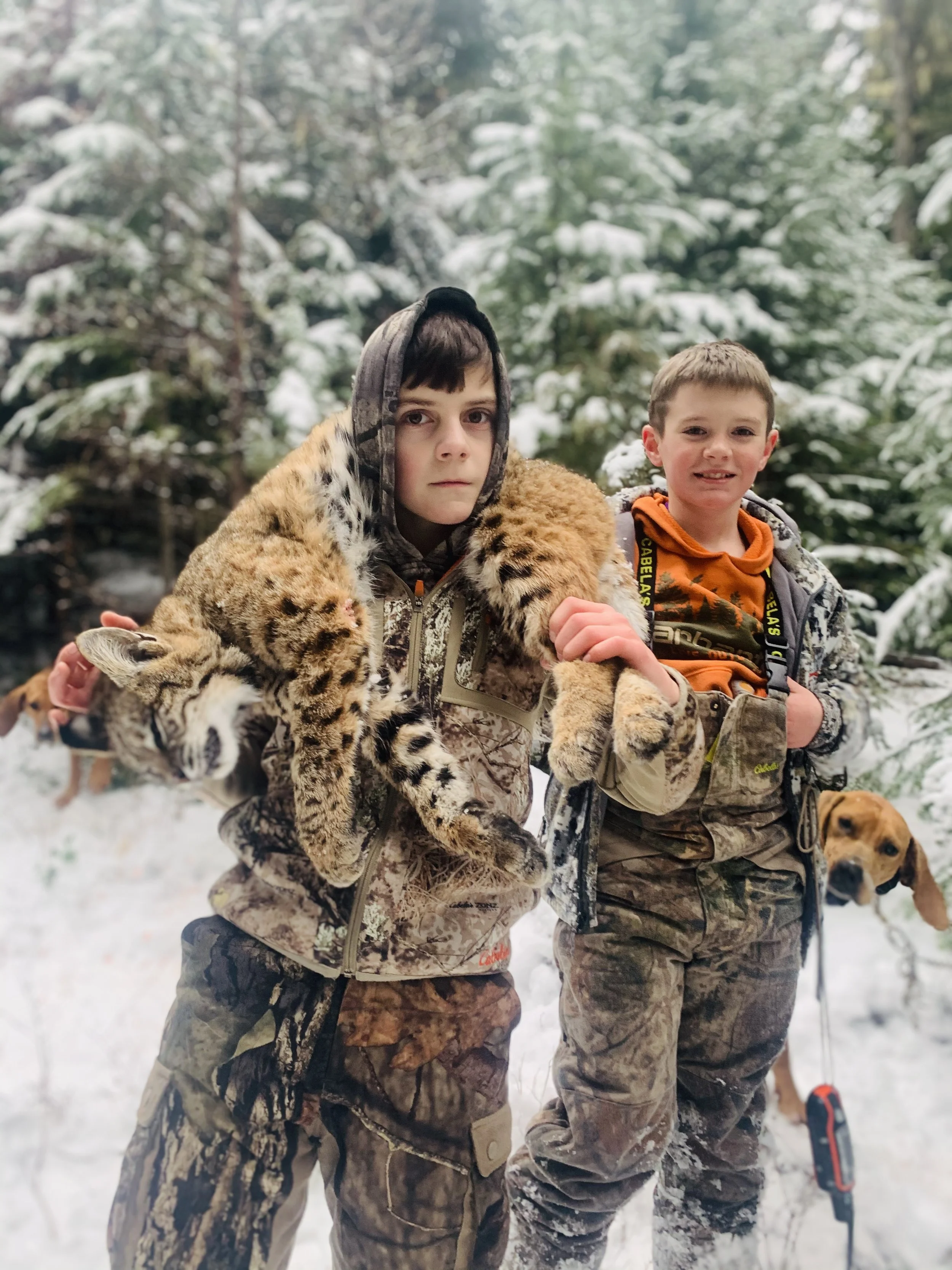 Two boys outdoors in snowy forest, one holding a young tiger and a dog in the background.