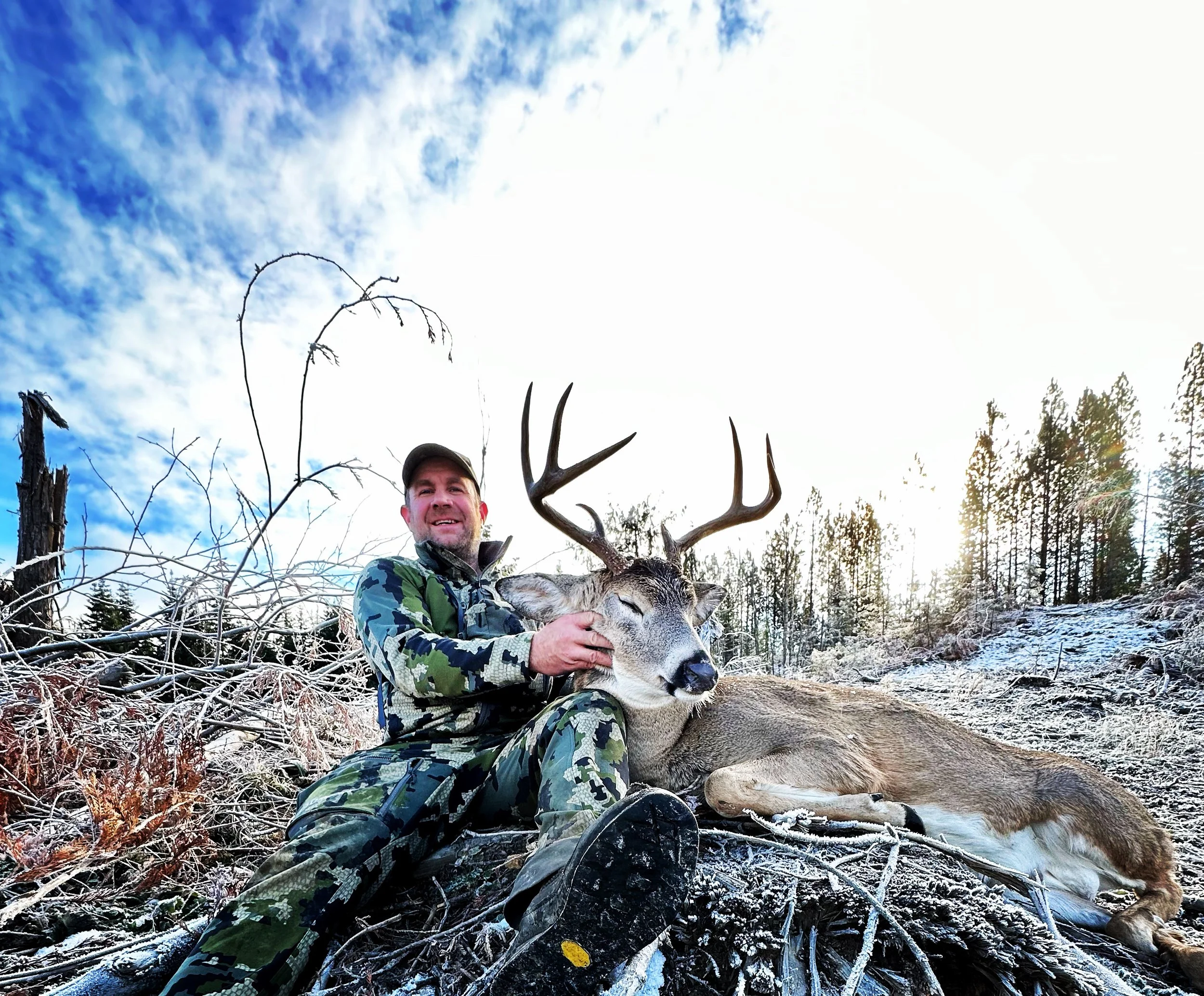 A man in camouflage clothing sitting outdoors in a snowy landscape, holding a large deer with antlers who is lying on the ground.