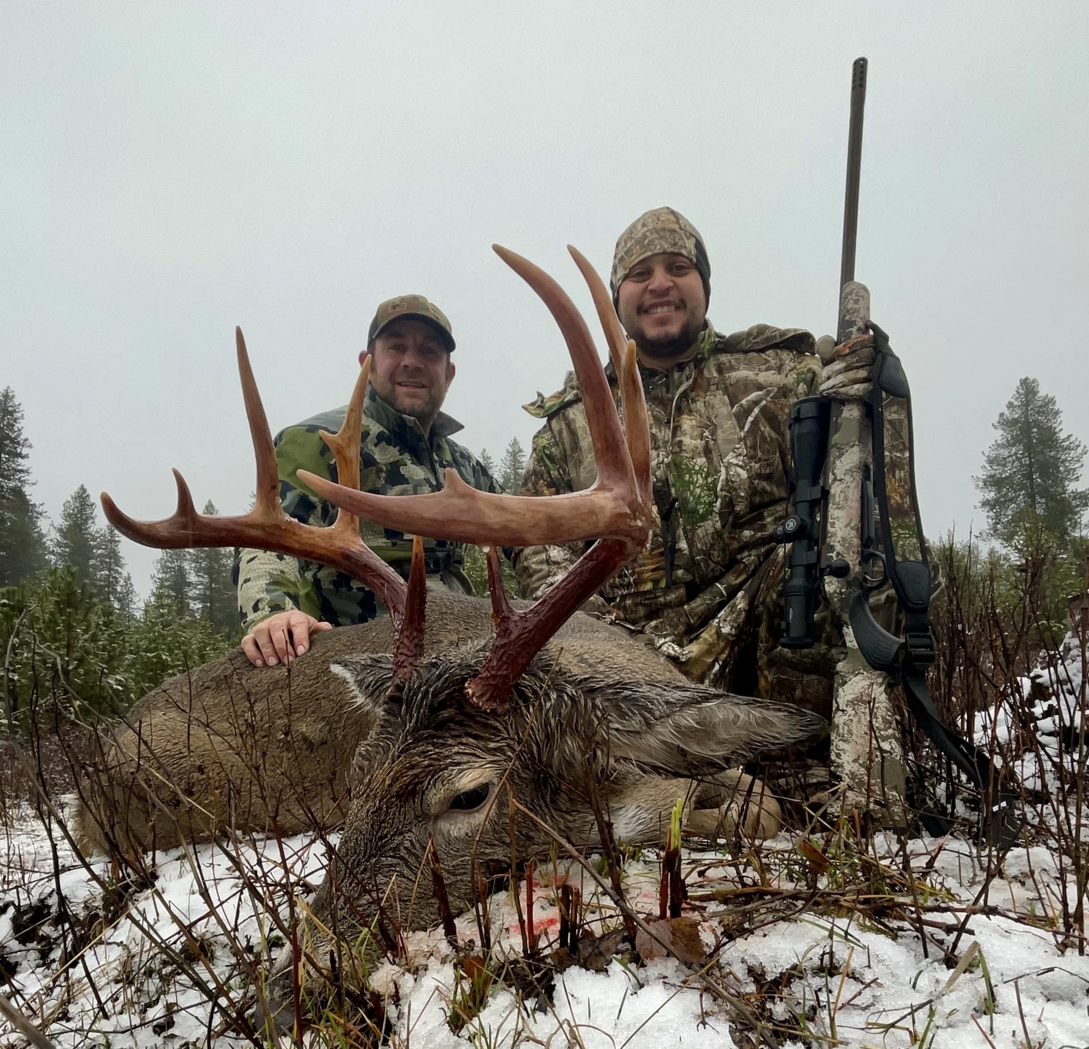 Two hunters in camouflage clothing posing with a large deer with antlers, lying on snowy ground in a forested area, one with a rifle.