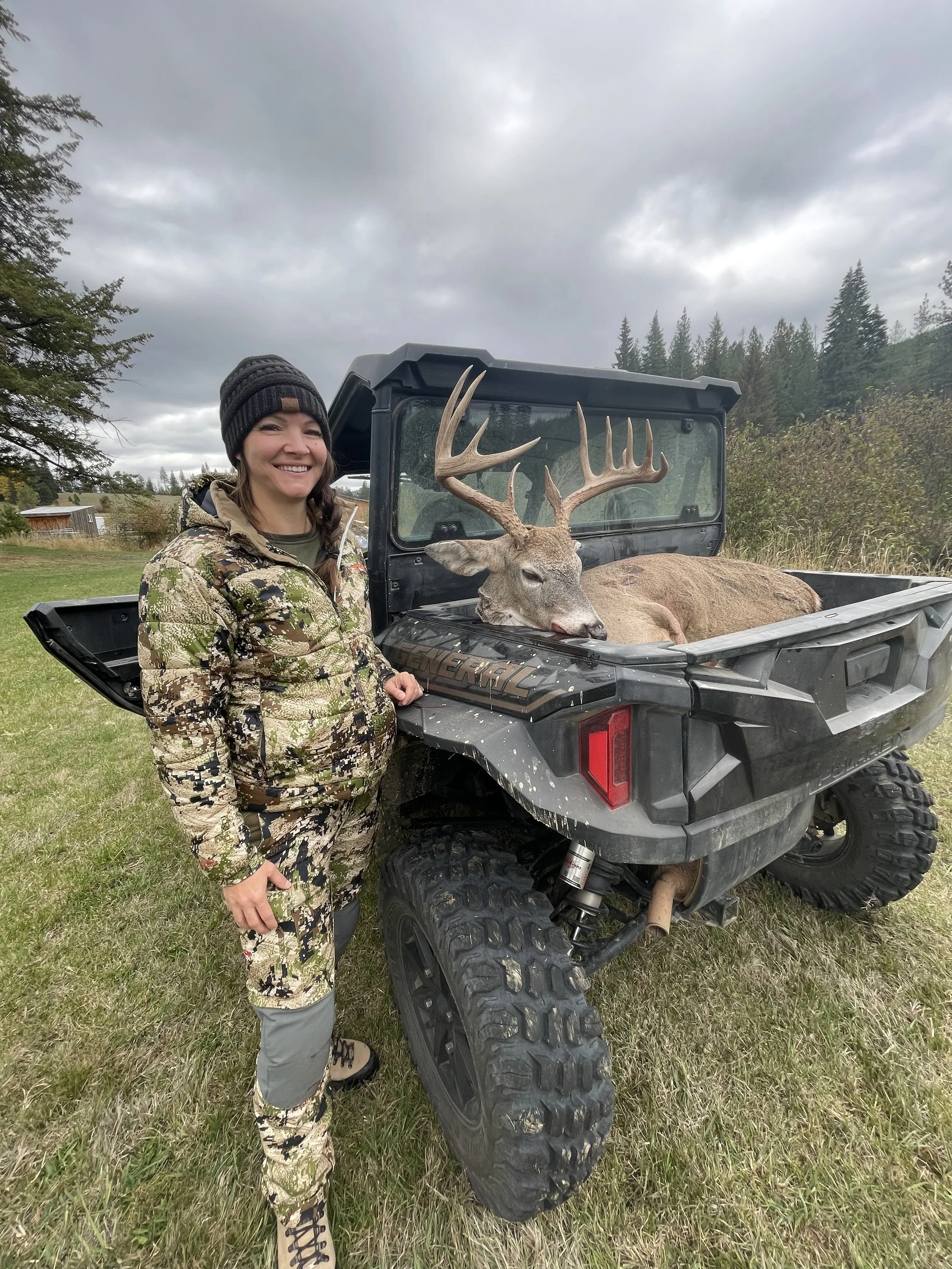 A woman in camouflage hunting gear standing next to an ATV with a large dead buck with antlers lying in the truck bed.