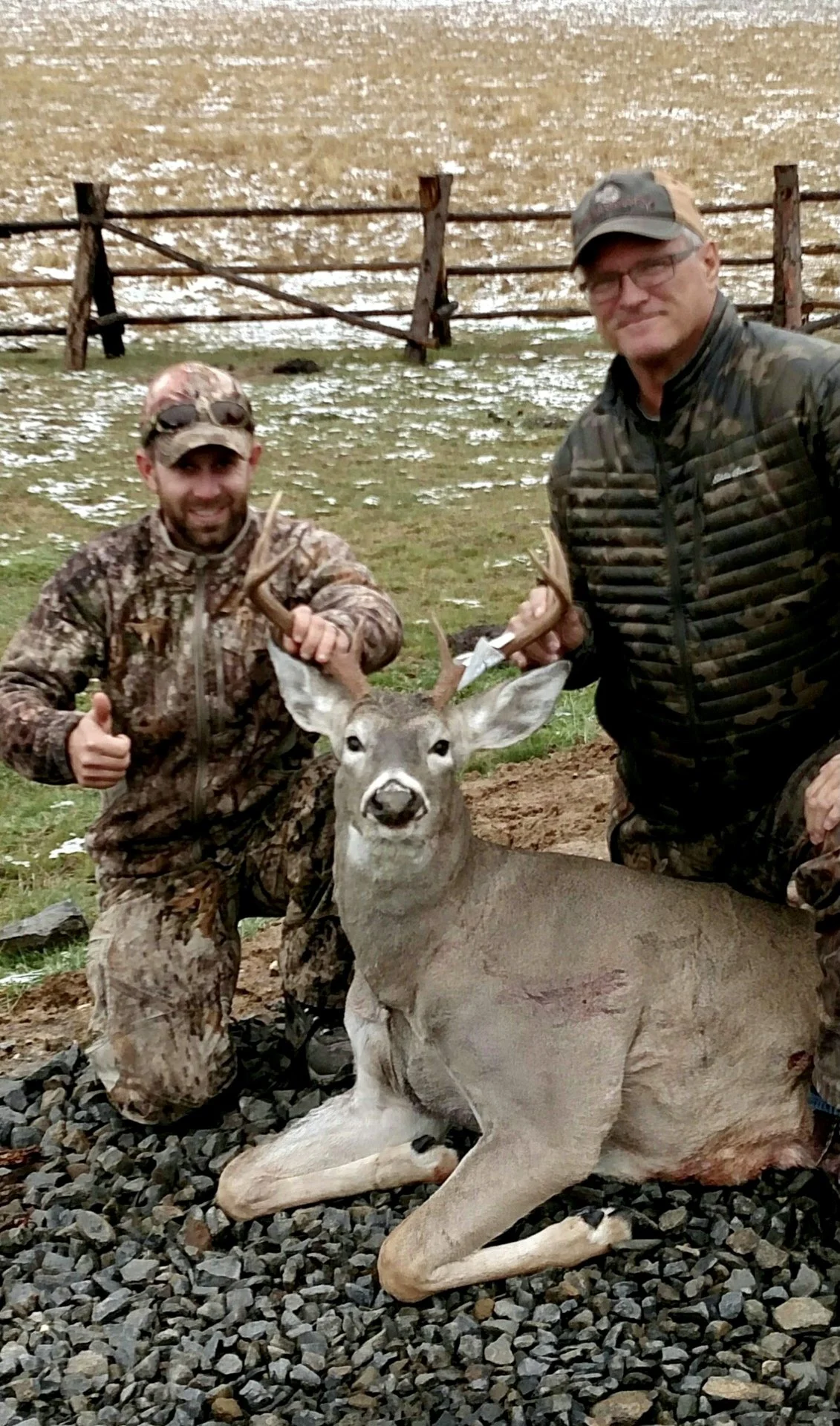 Two men in camouflage outdoor clothing kneel beside a freshly hunted deer with antlers, giving a thumbs-up and smiling outdoors on a rocky ground, with a wooden fence and field in the background.