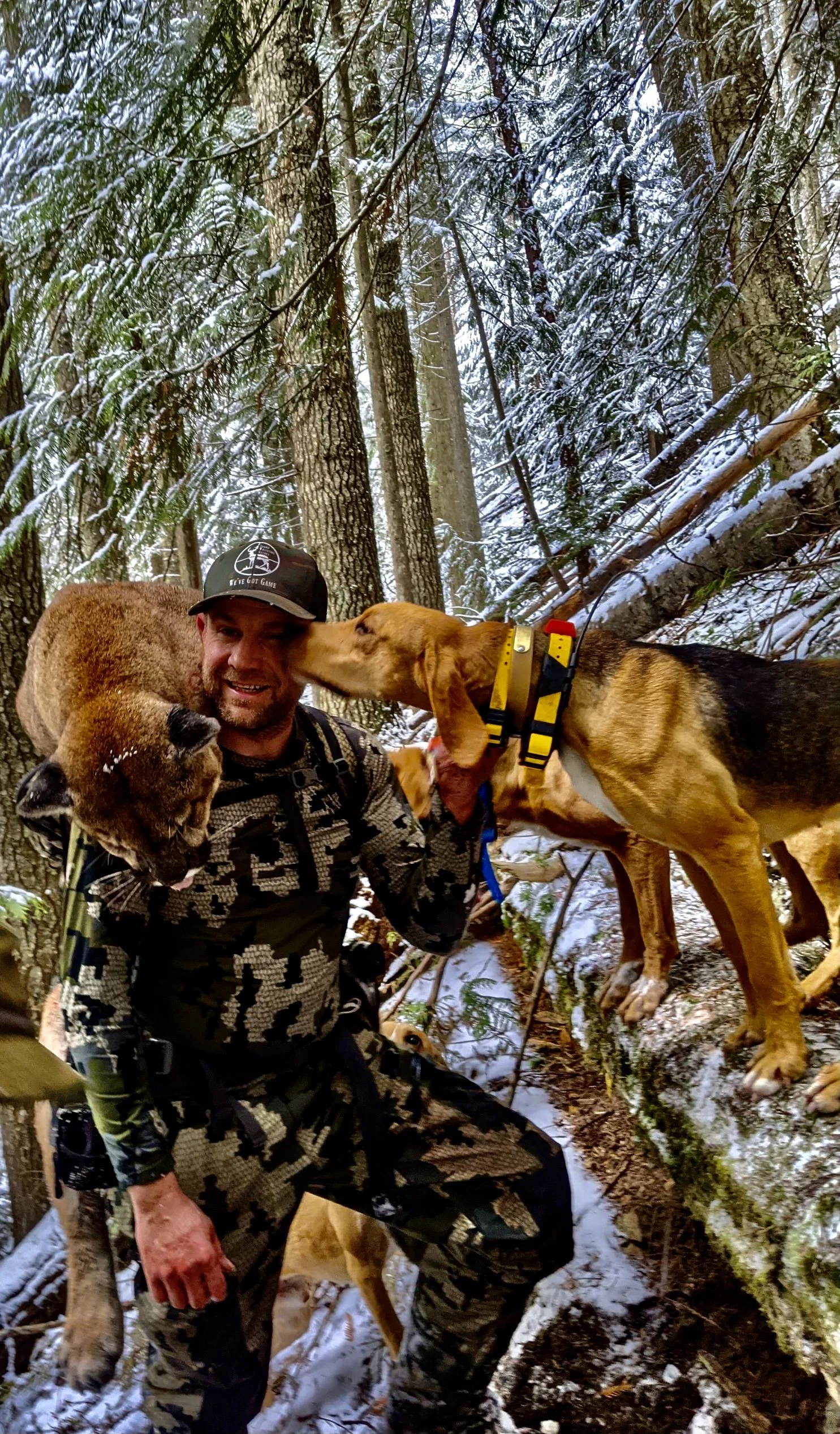 A man with a beard in camouflage clothes and a black cap kneels in a snow-covered forest, holding a large bear's head in his left hand and a bloodhound dog licking his face.