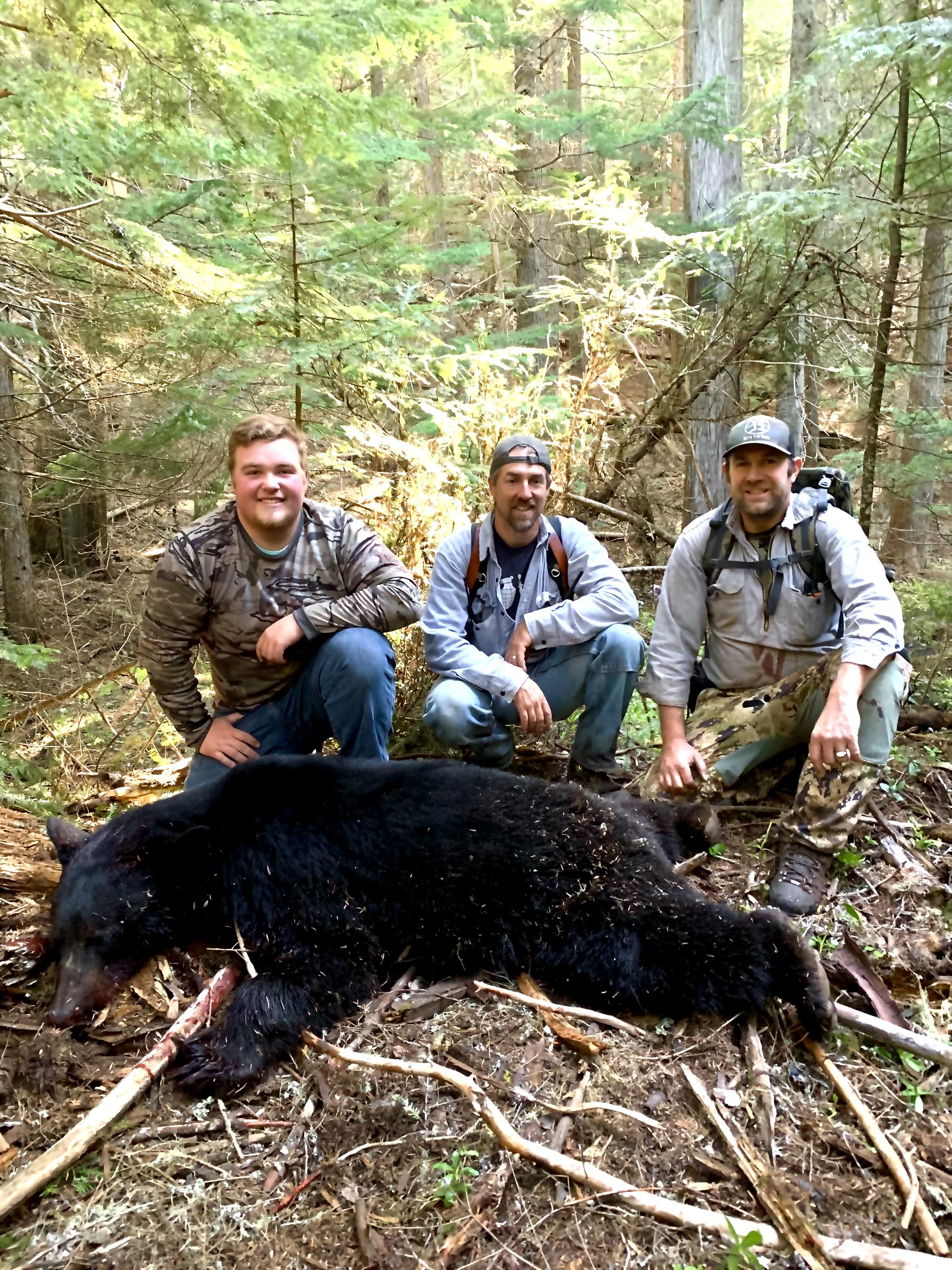 Three men crouching in a forested area with a large black bear carcass in front of them.