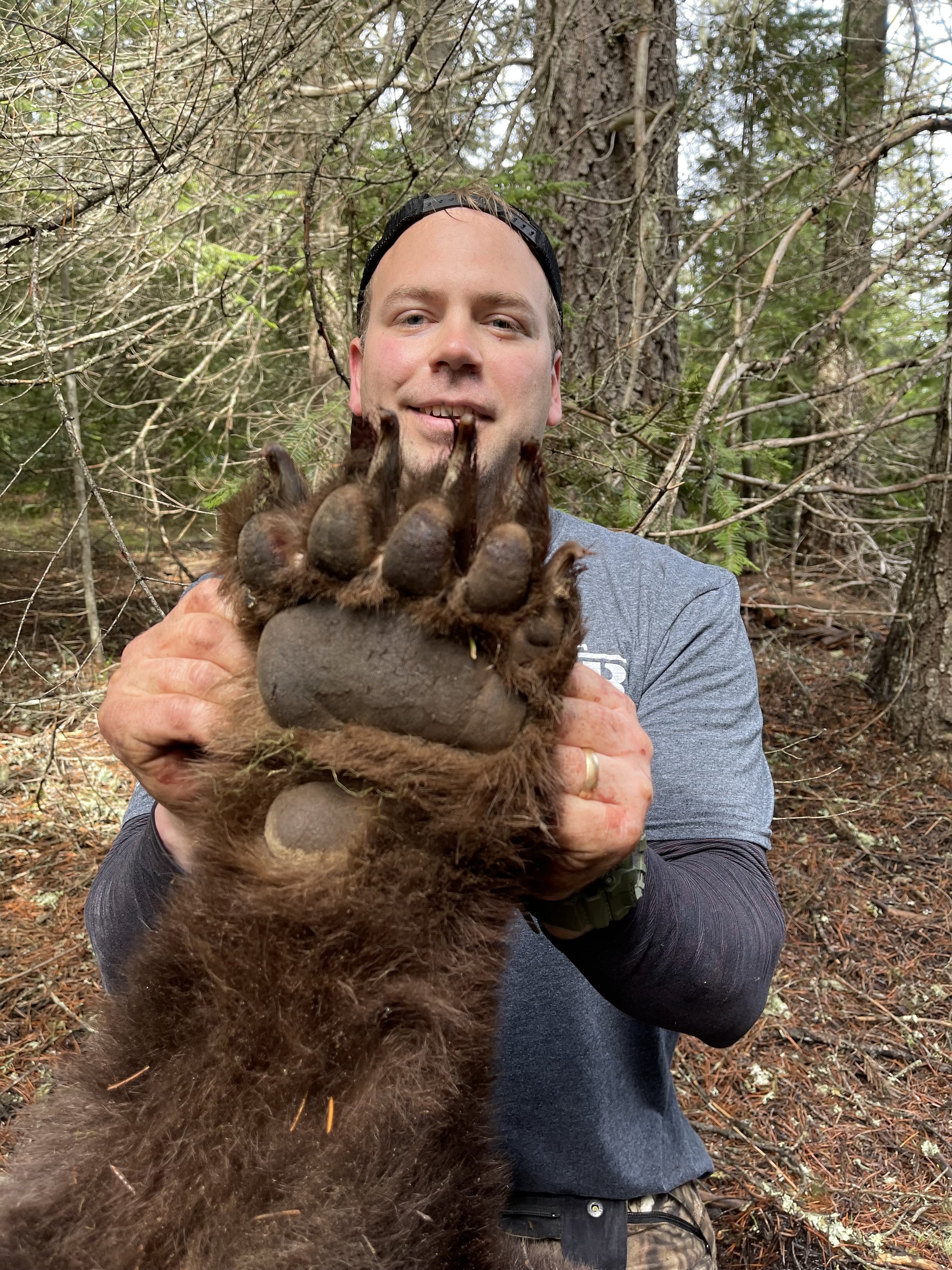 A man holding up a large, upside-down, fuzzy animal paw with claws in a forest. The man is smiling and wearing a gray shirt, watch, and a black cap backward. Trees and forest ground surround him.