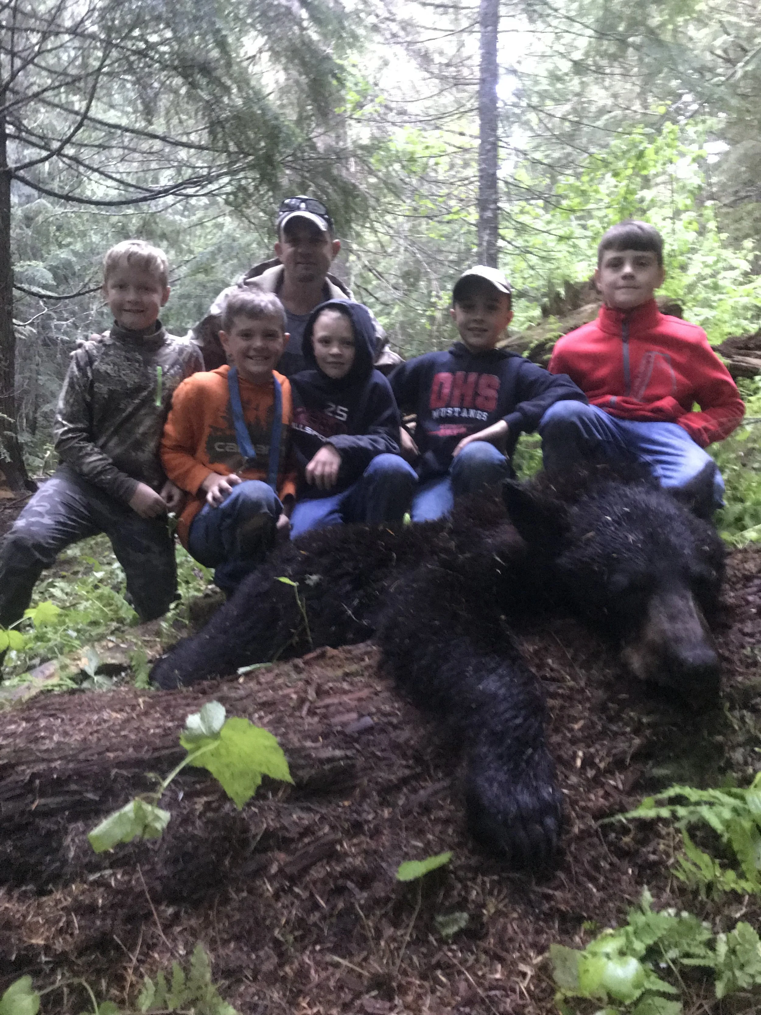 Six children and one adult pose behind a large dead bear in a forested area.