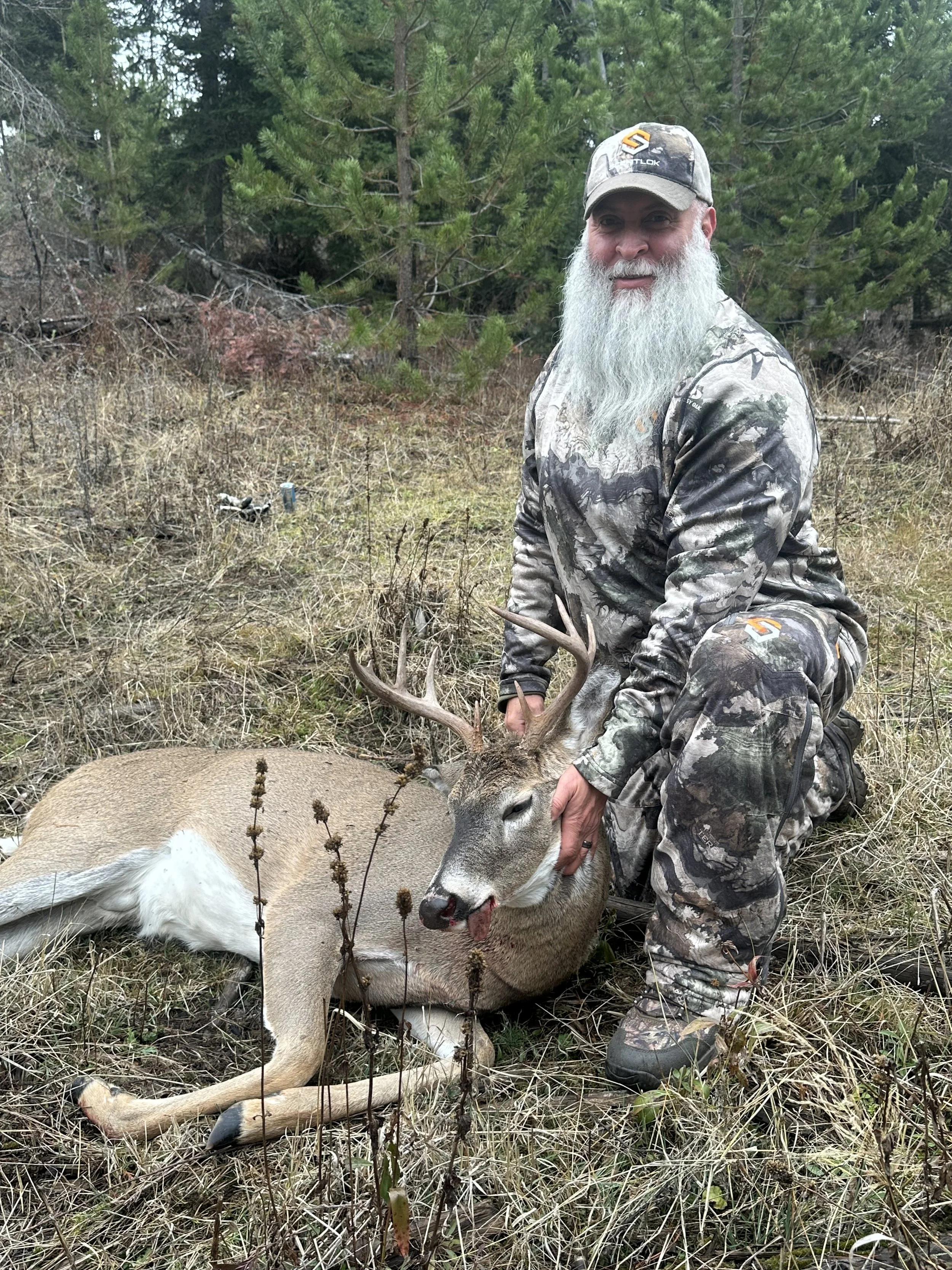 A man with a long white beard dressed in camouflage clothing kneels beside a freshly hunted deer with antlers, holding its head in a grassy forest area.