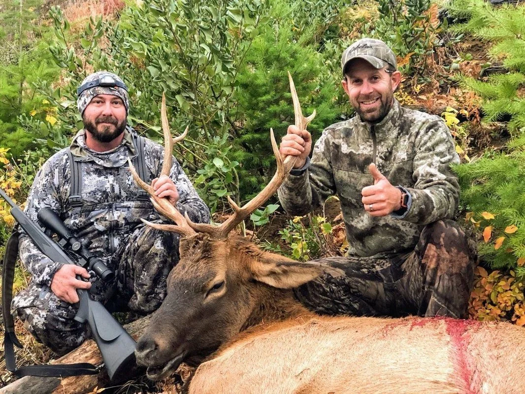 Two hunters in camouflage clothing posing with a large deer they have hunted, outdoors with green foliage in the background.