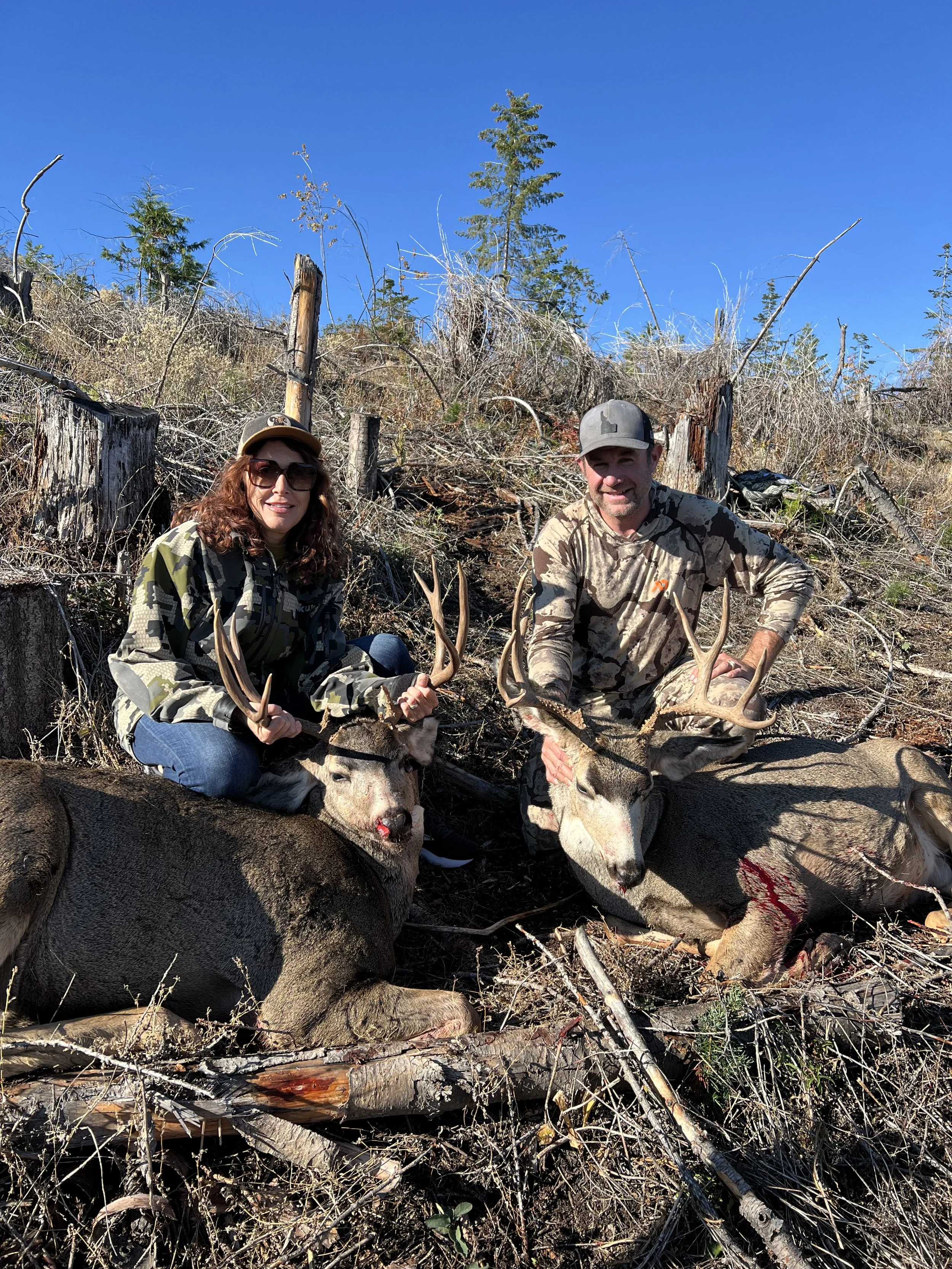 Two hunters, a woman and a man, pose with large harvested deer in a forested area. The woman holds one deer’s antlers and is smiling, wearing sunglasses and camouflage clothing. The man is kneeling next to another deer, holding its antlers and smilin
