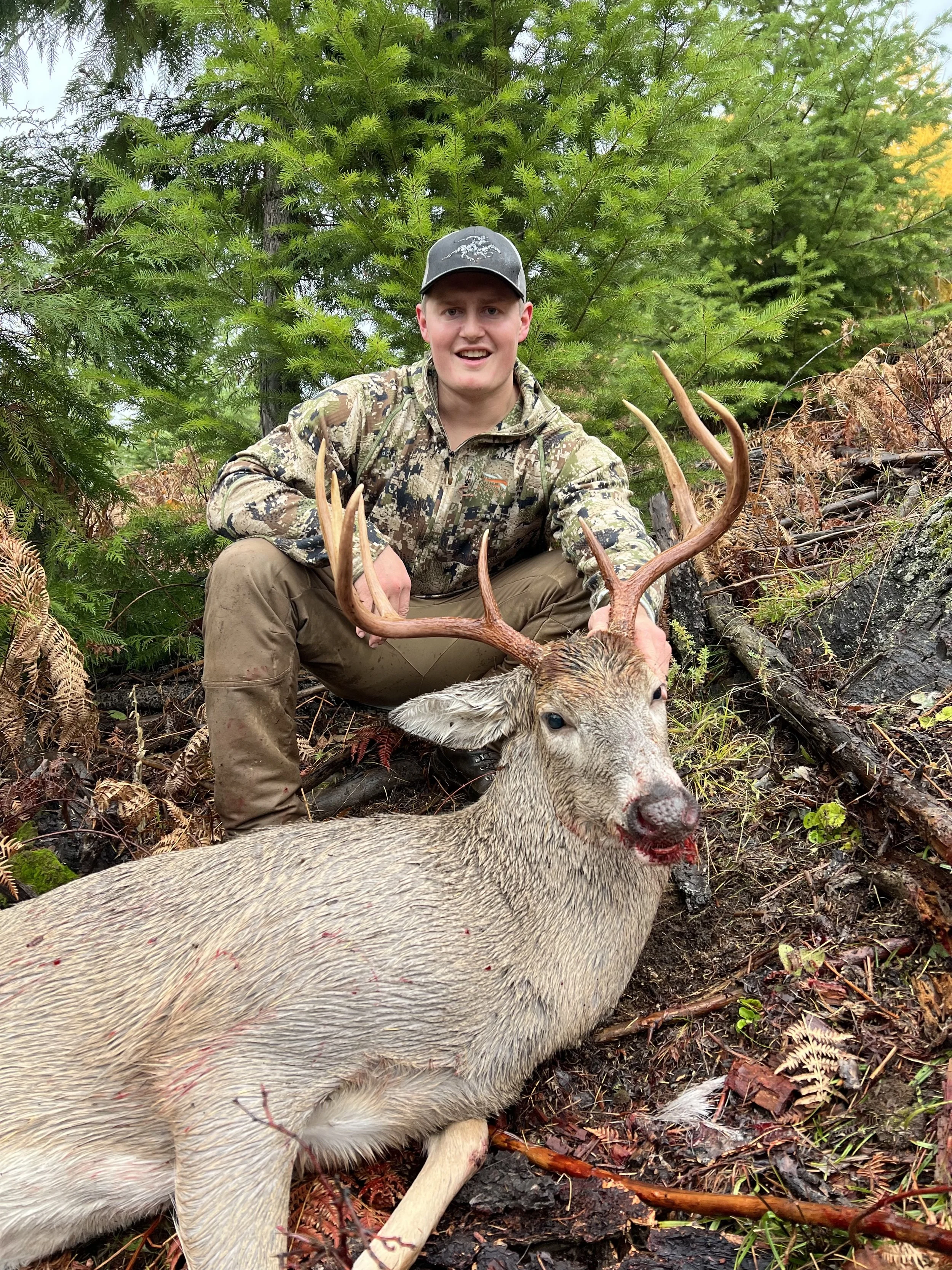 A young man in camouflage clothing and a baseball cap crouches next to a deceased deer with large antlers in a forested area, with green trees and foliage in the background.