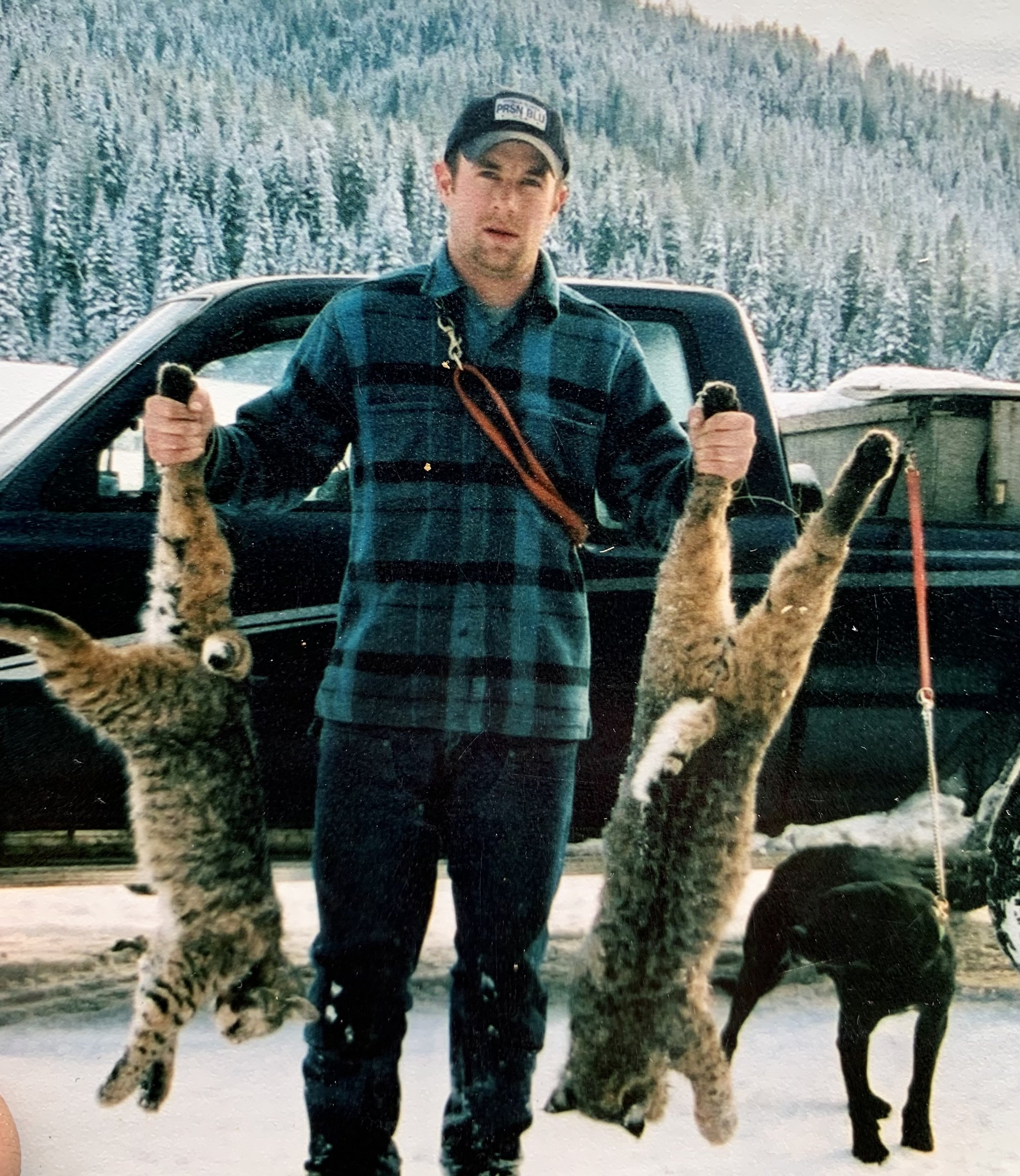 A man holding two bobcats by their front paws, standing in a snowy outdoor area with a dark vehicle and snow-covered trees in the background.
