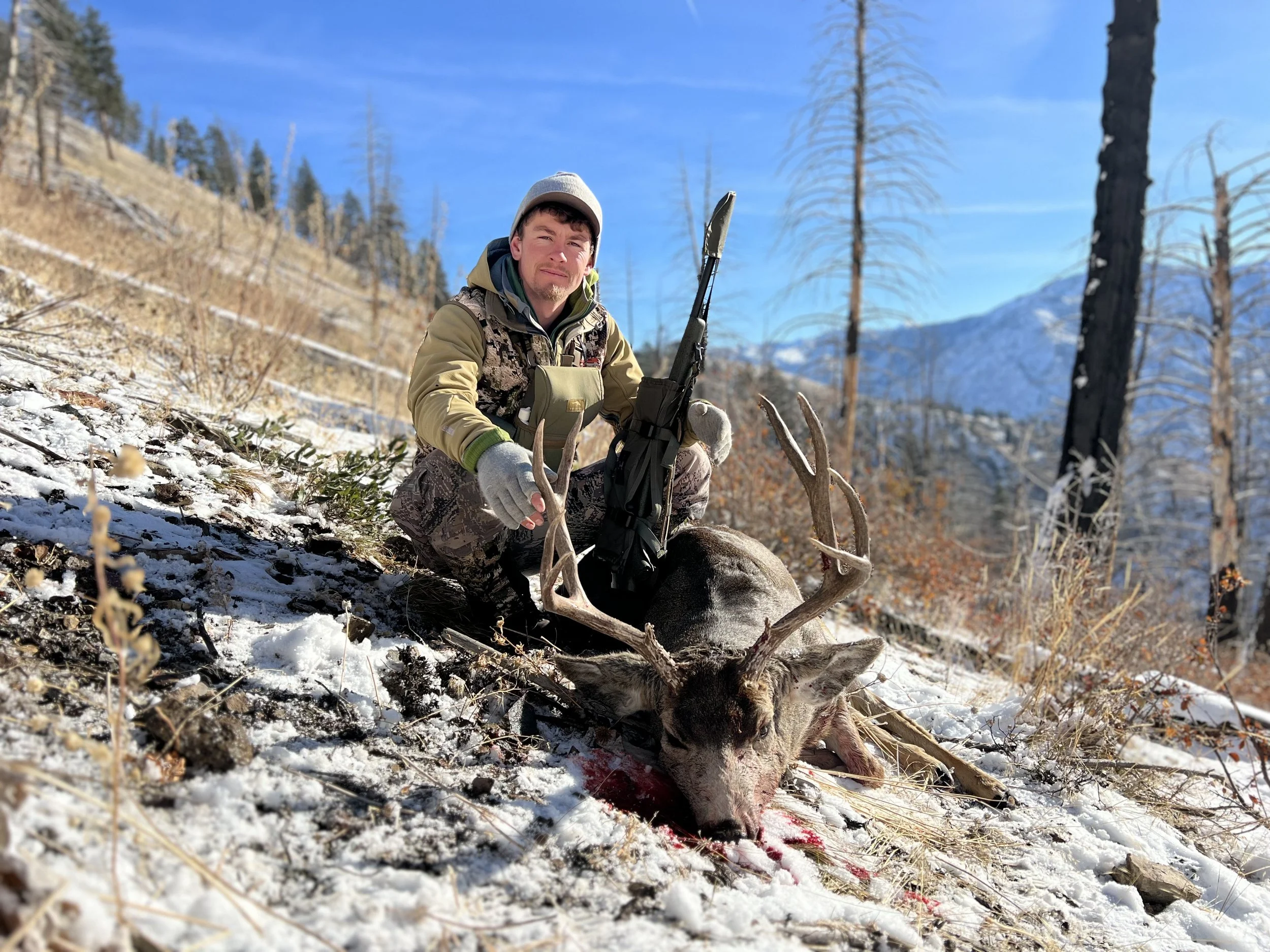 A man in camouflage clothing squats next to a deer with large antlers in a snowy forested area, holding a rifle in the ground.