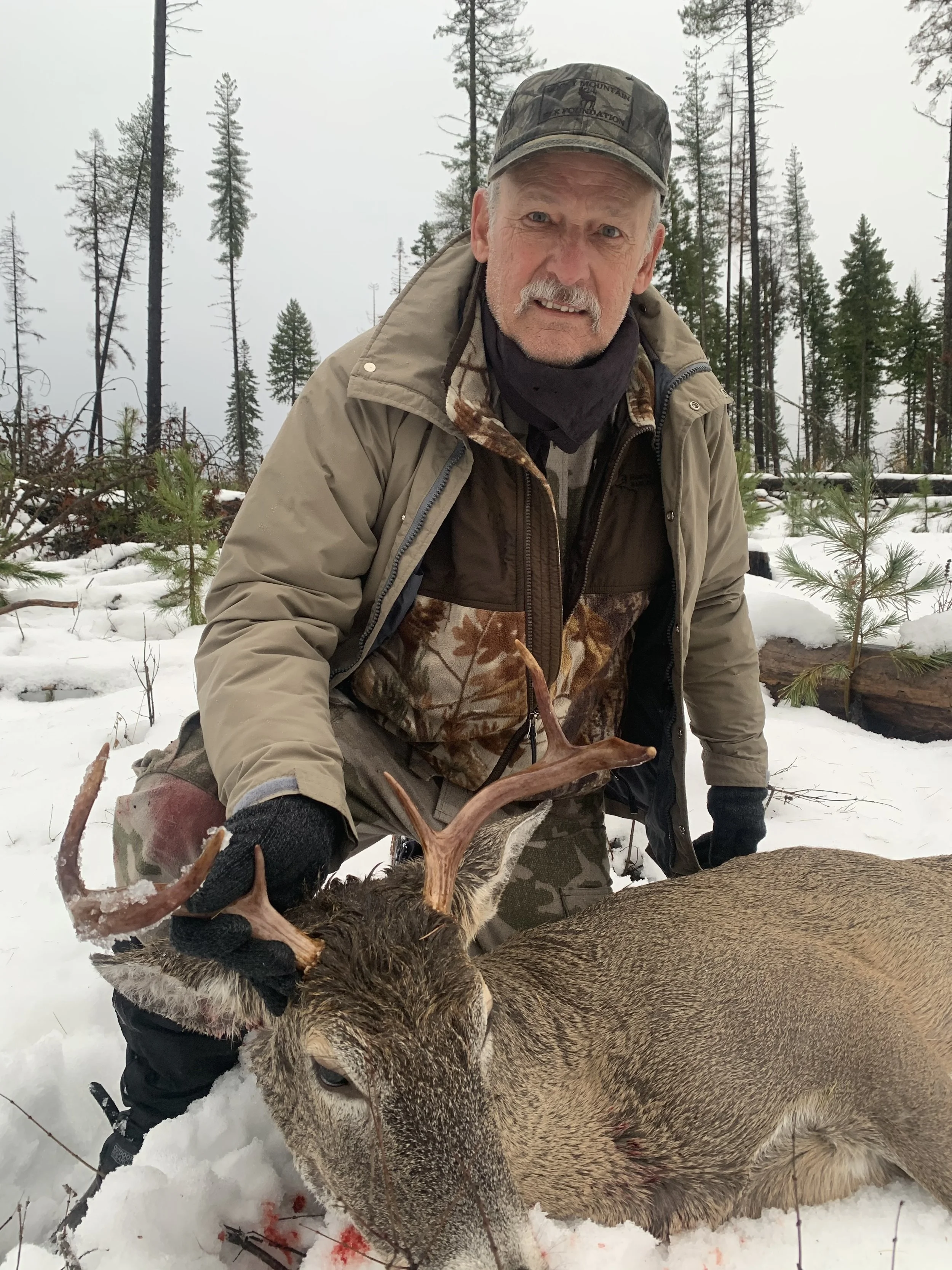 A man kneeling in the snow holding the antlers of a dead deer, with a forested background and snow-covered ground.