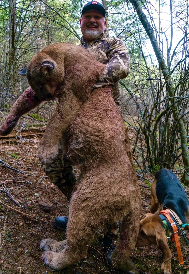 A man in camouflage clothing holding a large dead bear in a forest, with a dog nearby.