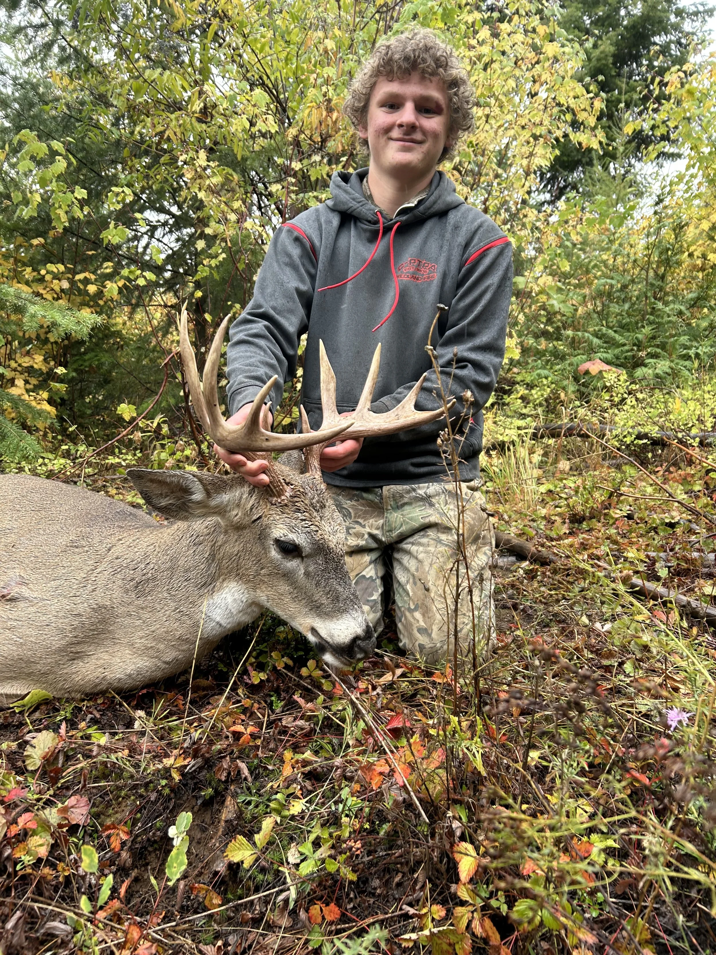 A young man in a hoodie and camouflage pants kneels next to a large buck deer with antlers in a wooded area, holding the antlers.