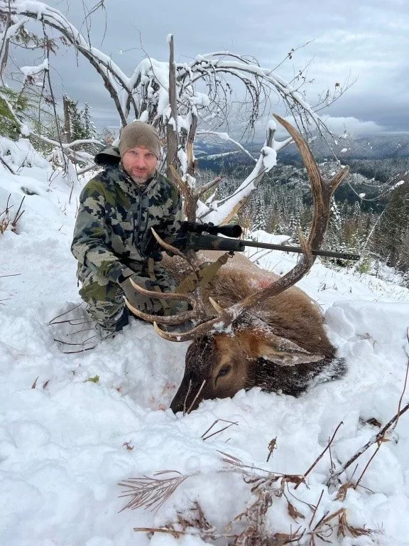 A man in camouflage clothing kneeling next to a large dead elk with antlers in a snowy outdoor setting.