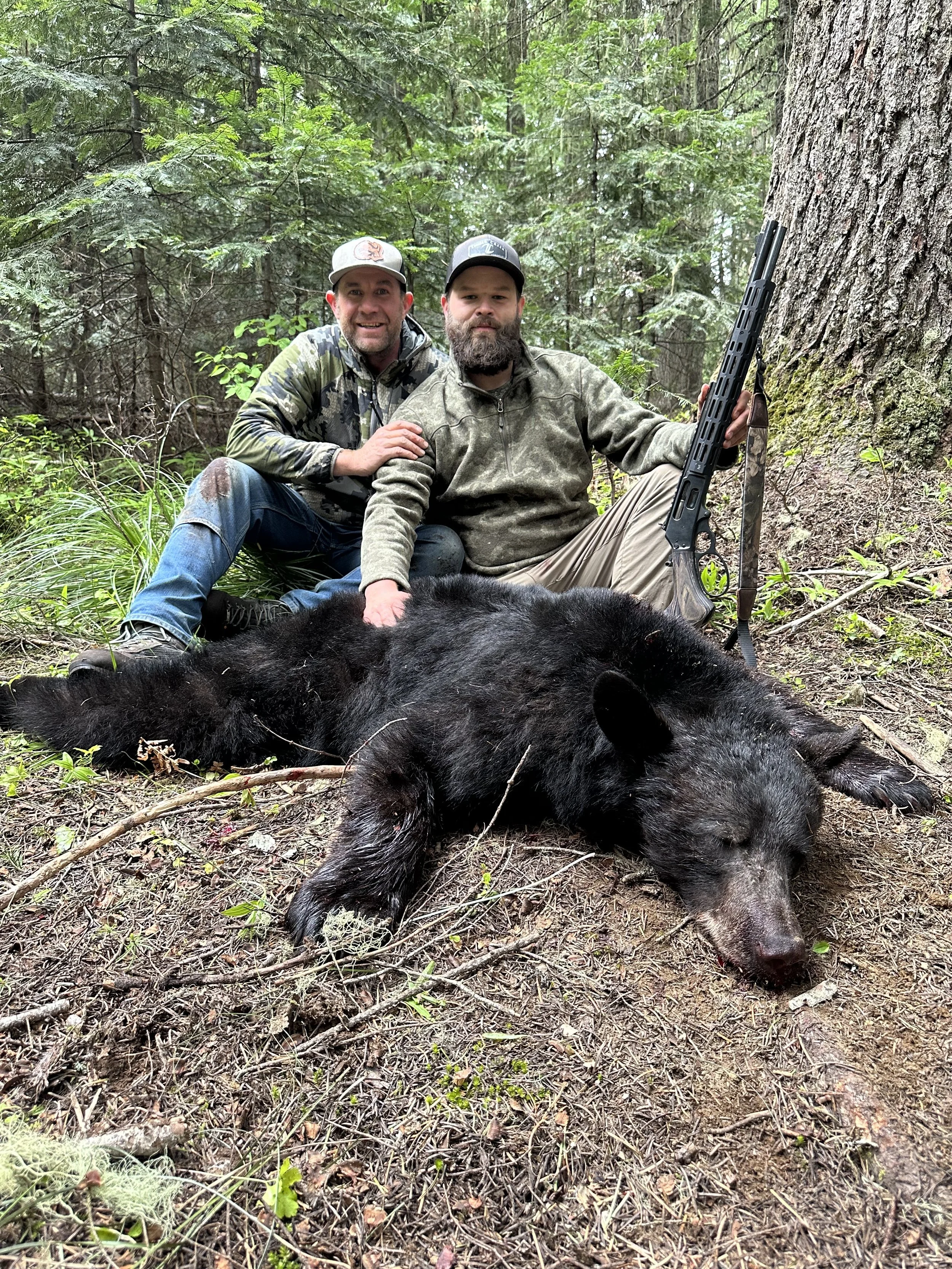 Two men in camouflage and outdoor clothing are sitting next to a large black bear that is lying on the forest ground, with a rifle leaning against a tree nearby.