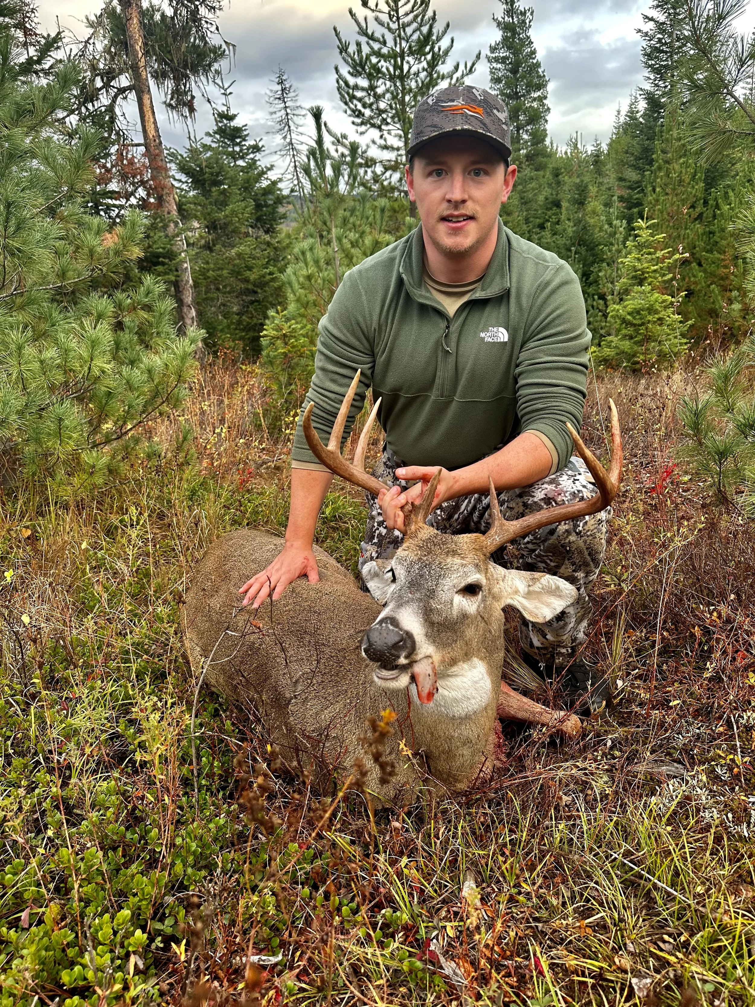 A man kneeling in a forested area with pine trees, holding the antlers of a large deer with a big set of antlers, showing the deer lying on the ground with blood on its nose and tongue.