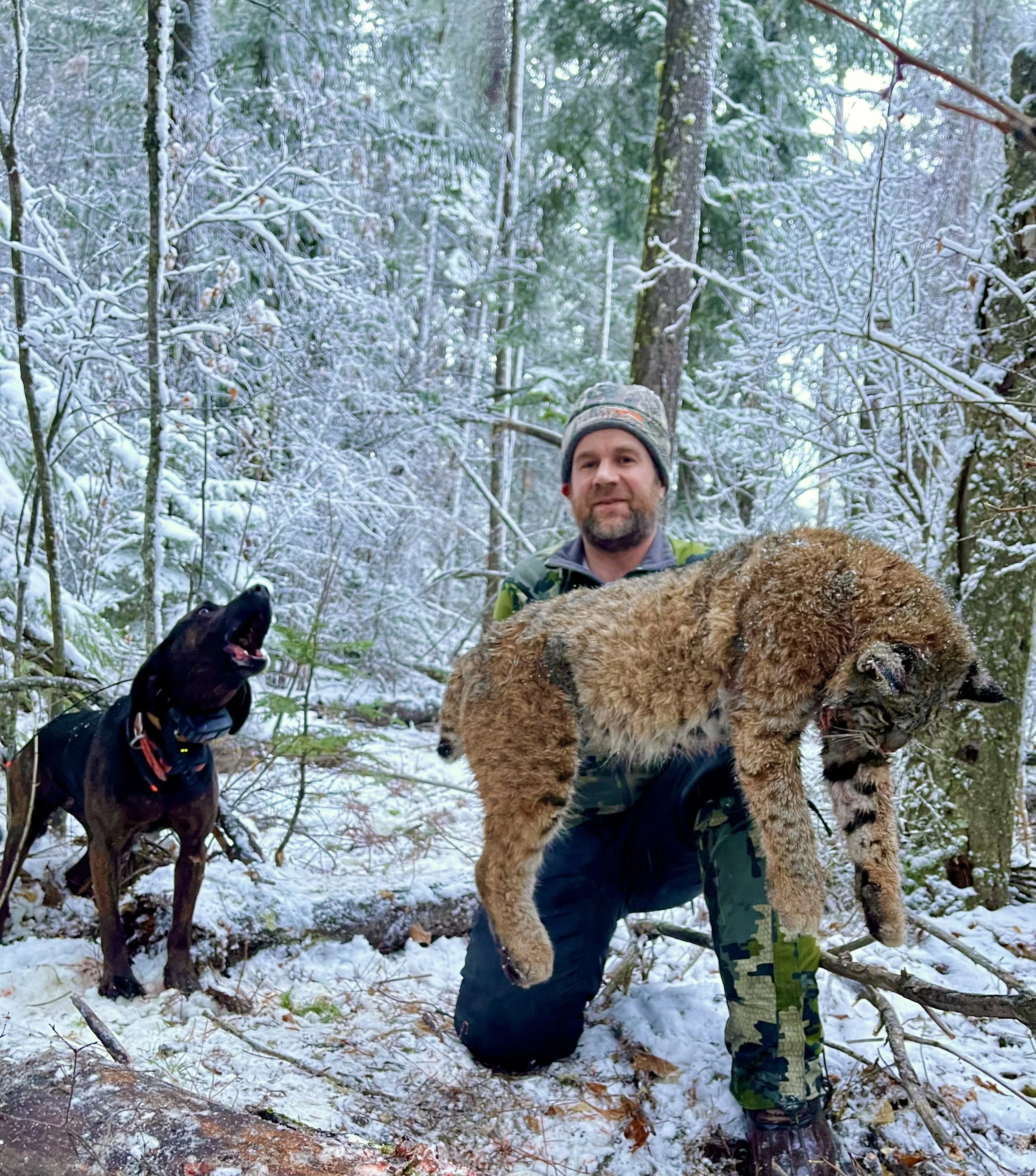 A man kneeling in a snowy forest holding a captured animal, possibly a young bear, with a black dog standing nearby.