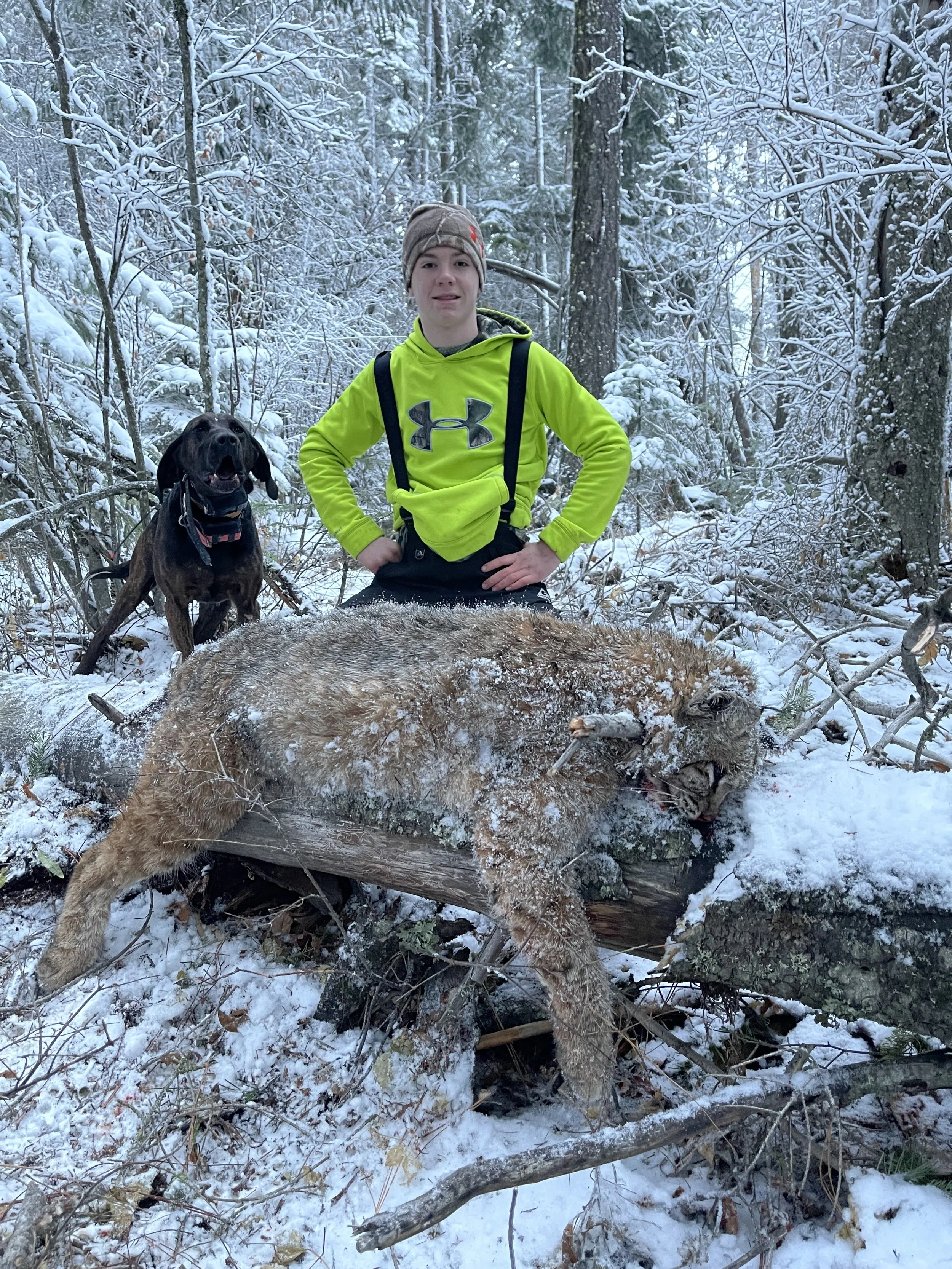 A young boy wearing a bright yellow jacket and camouflage hat in a snow-covered forest, standing behind a large snow-covered animal, possibly a mountain lion, that is lying on a fallen tree. A dog stands nearby in the background.
