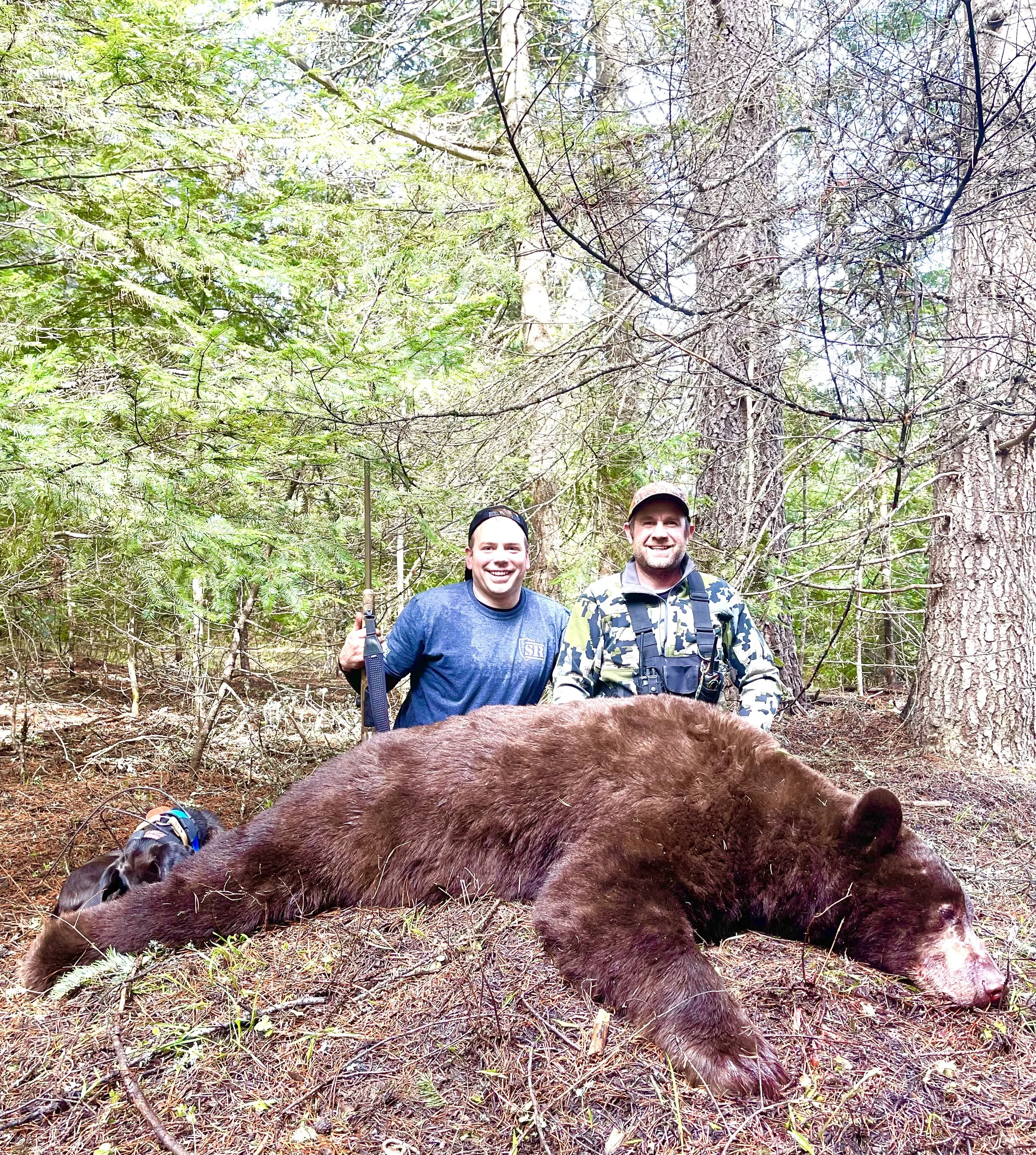 Two smiling hunters standing in a forest behind a large, dead bear lying on the ground.