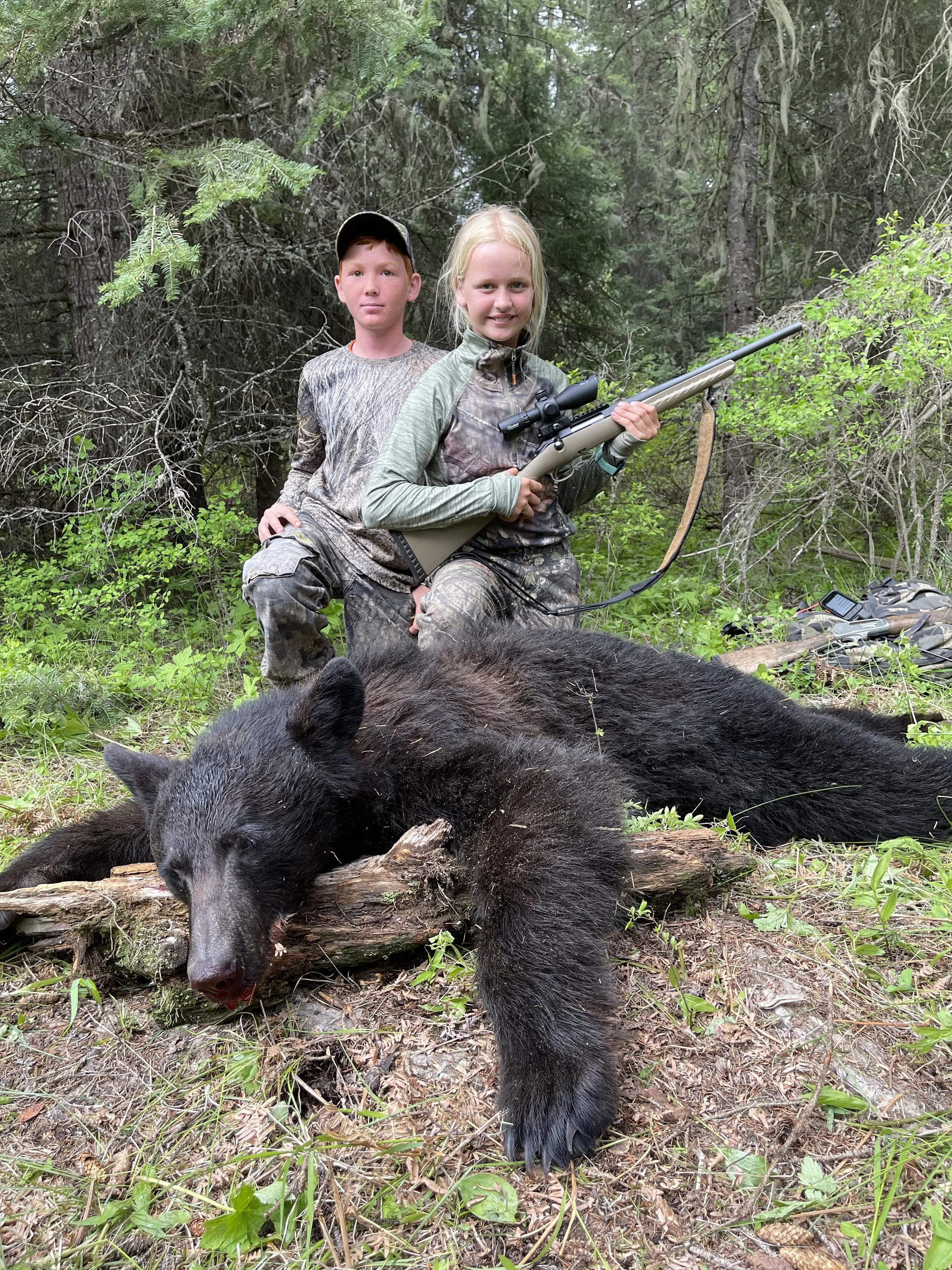 A girl holding a rifle and a boy behind her, both dressed in camouflage, stand in a forested area next to a large dead black bear lying on the ground.
