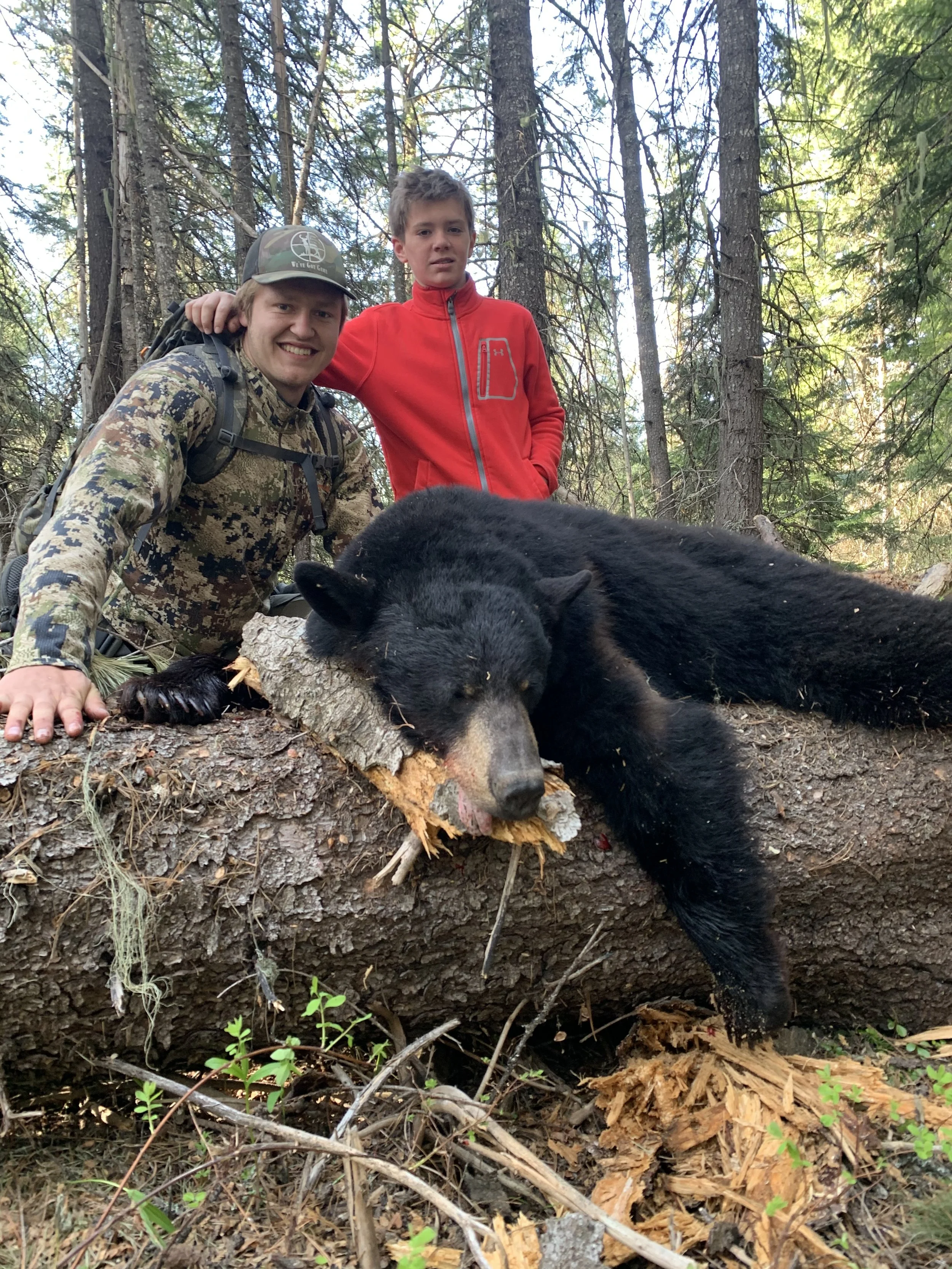 Two people in a forested area with a dead black bear on a fallen tree log, one person, in a camouflage shirt and cap, is smiling and crouching with one hand on the log, while the other person, in a red jacket, stands with their hand on the first pers