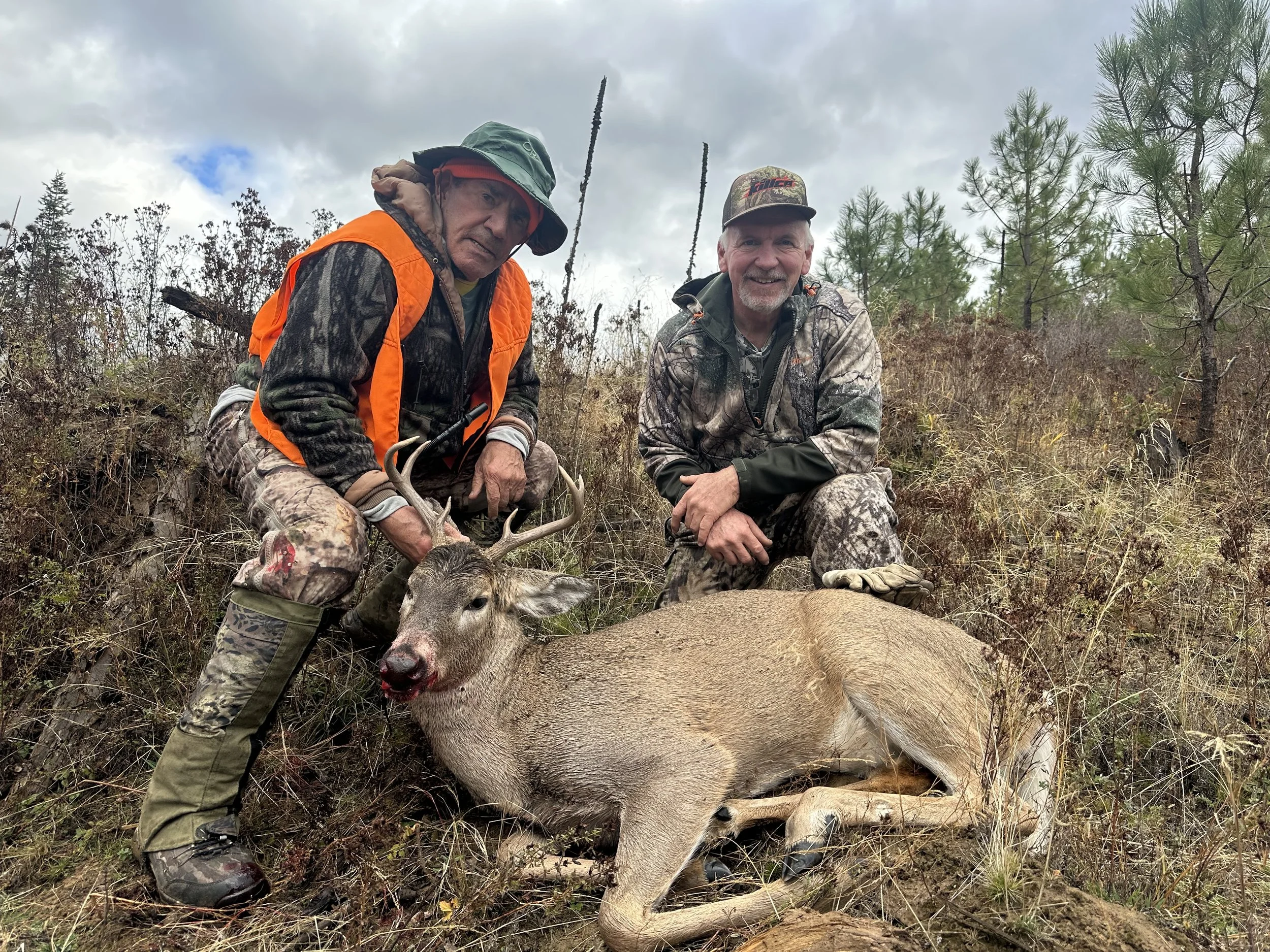 Two hunters in camouflage and orange safety gear kneeling next to a freshly hunted deer with antlers in a natural outdoor setting with trees and cloudy sky.