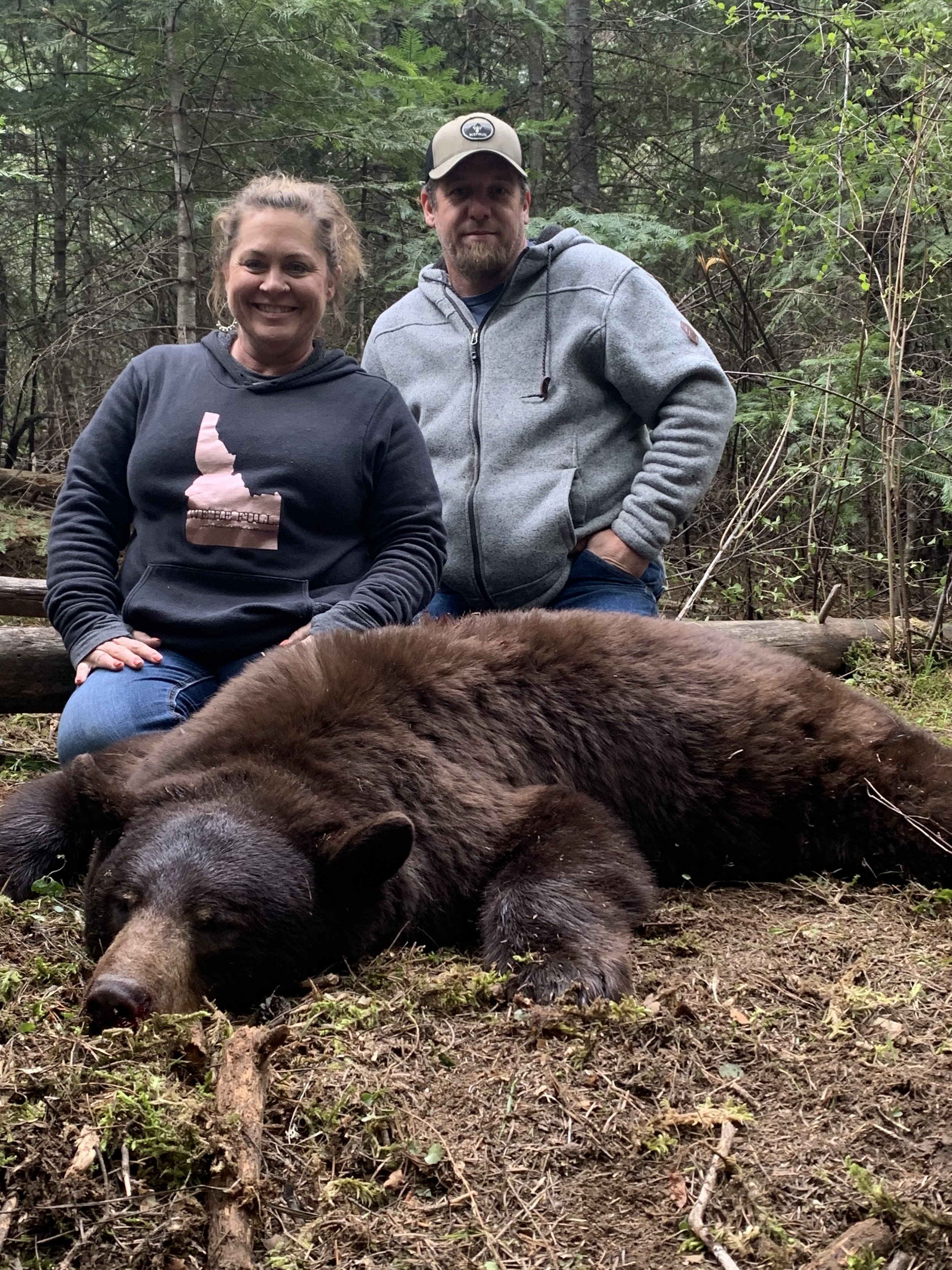 A woman and a man kneeling behind a large, sleeping brown bear in a forest setting.