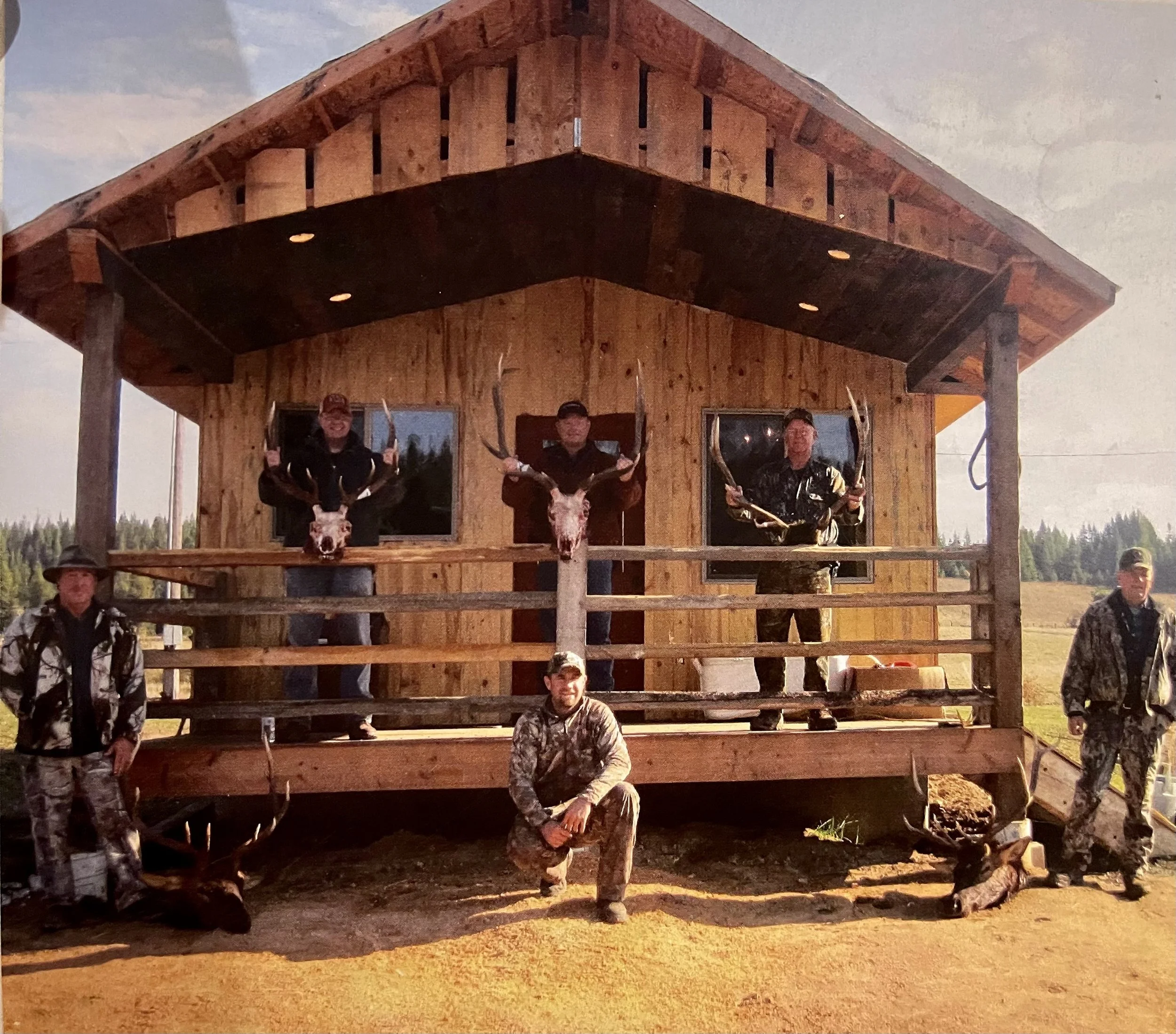 Group of men displaying hunted deer and antlers in front of a wooden cabin in a rural outdoor setting.