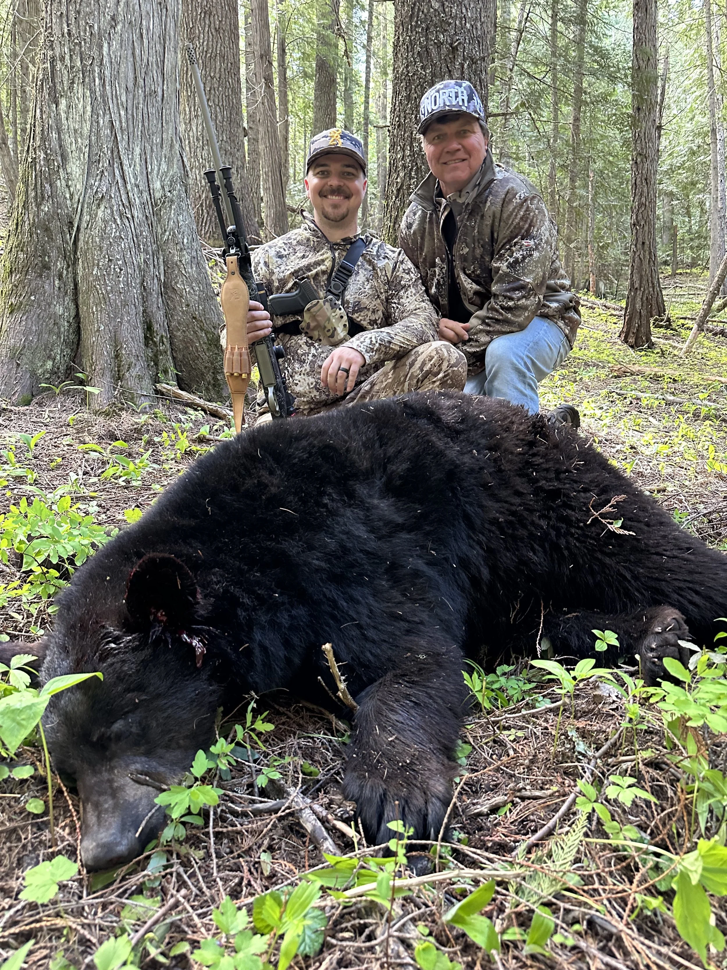 Two men in camouflage clothing posing in the woods with a dead black bear in front of them, one holding a rifle.