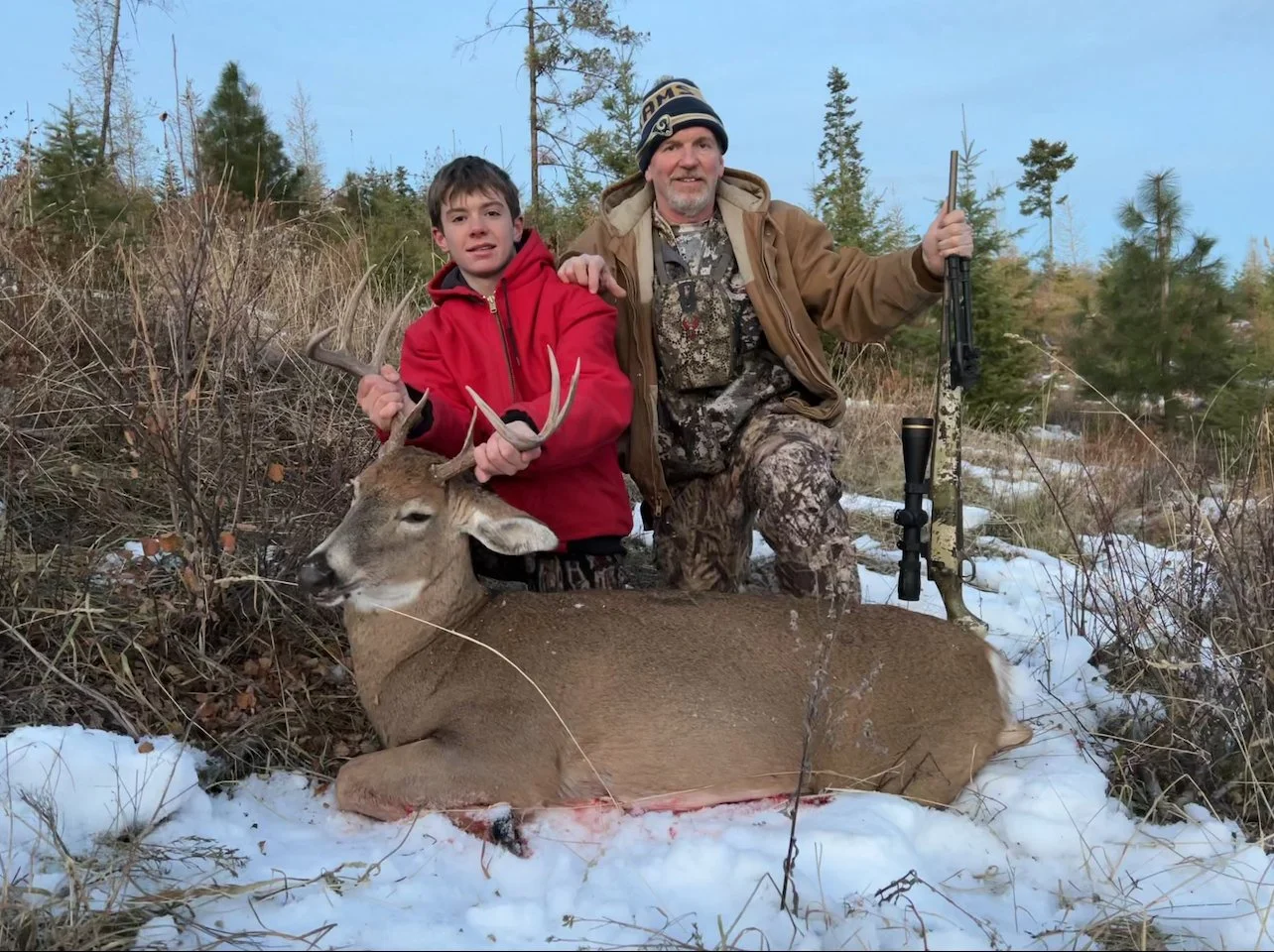 A boy and a man outdoors in a snowy area with trees, holding a deer they have hunted, with the boy holding the antlers and the man holding a rifle.