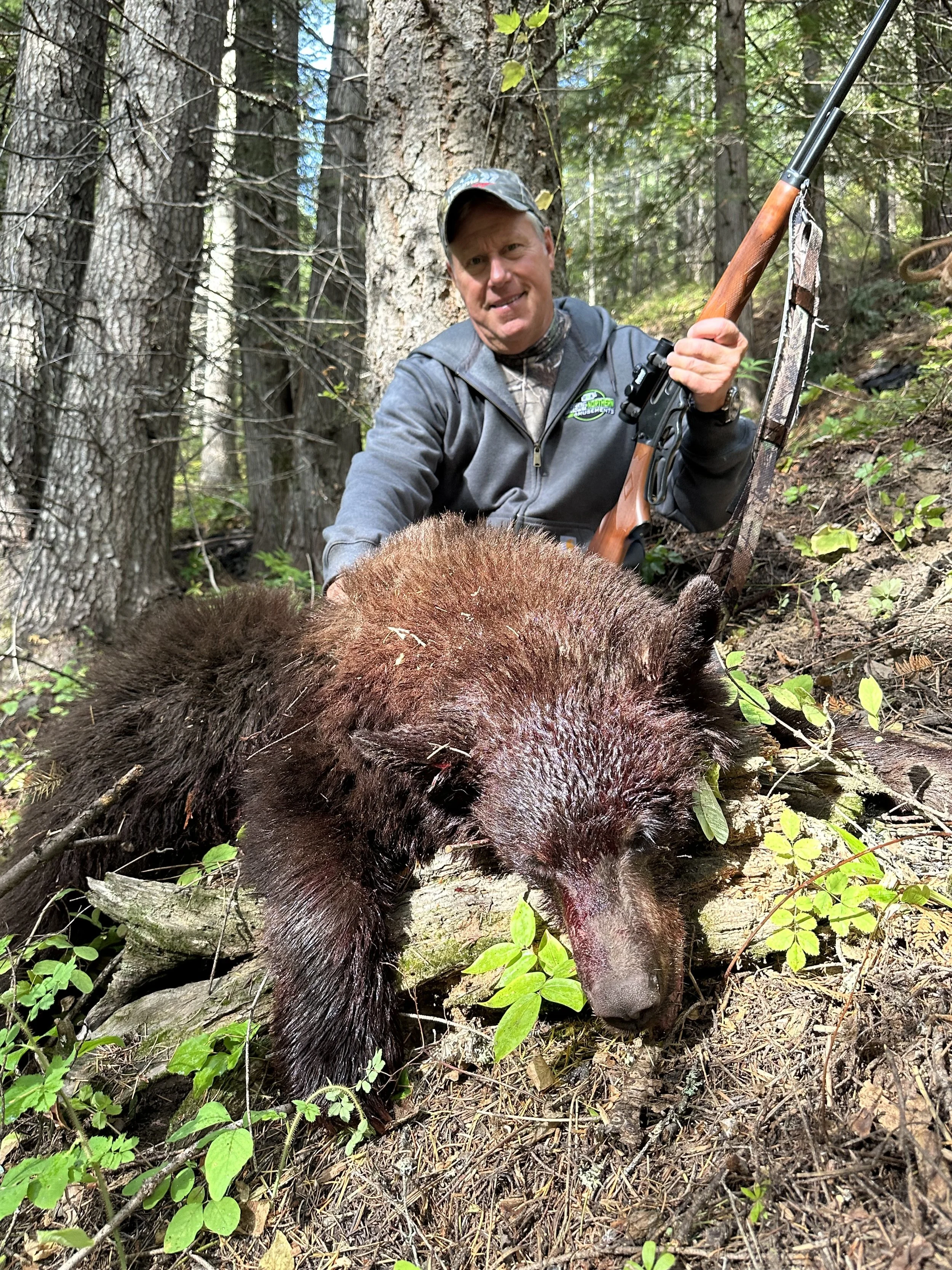 A man in outdoor gear kneeling in a forested area with a rifle, posing behind a deceased bear lying on the ground among trees and green foliage.