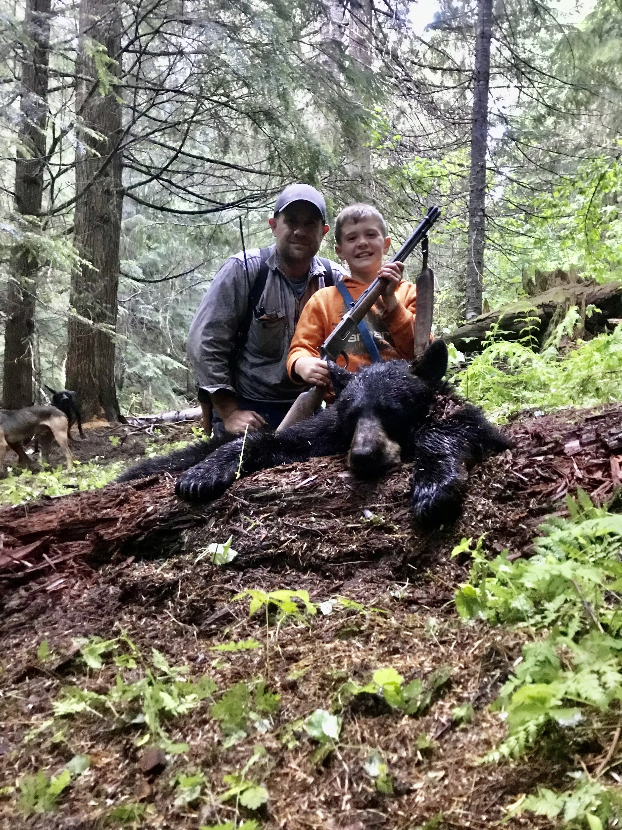 A man and a boy are posing in a forest with a dead black bear lying in front of them. The boy is holding a rifle and a large fish. The forest is dense with green foliage. There are other animals, including a dog, in the background.