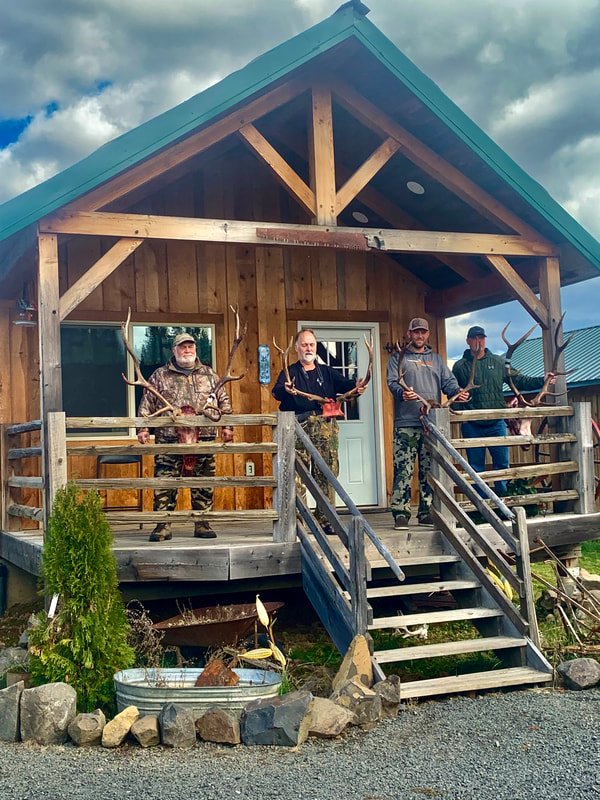 Four men standing on a porch of a wooden cabin, each holding a large antlered game animal. The cabin has a porch with stairs leading up and a green roof. There are rocks, plants, and a small pond in front of the cabin.