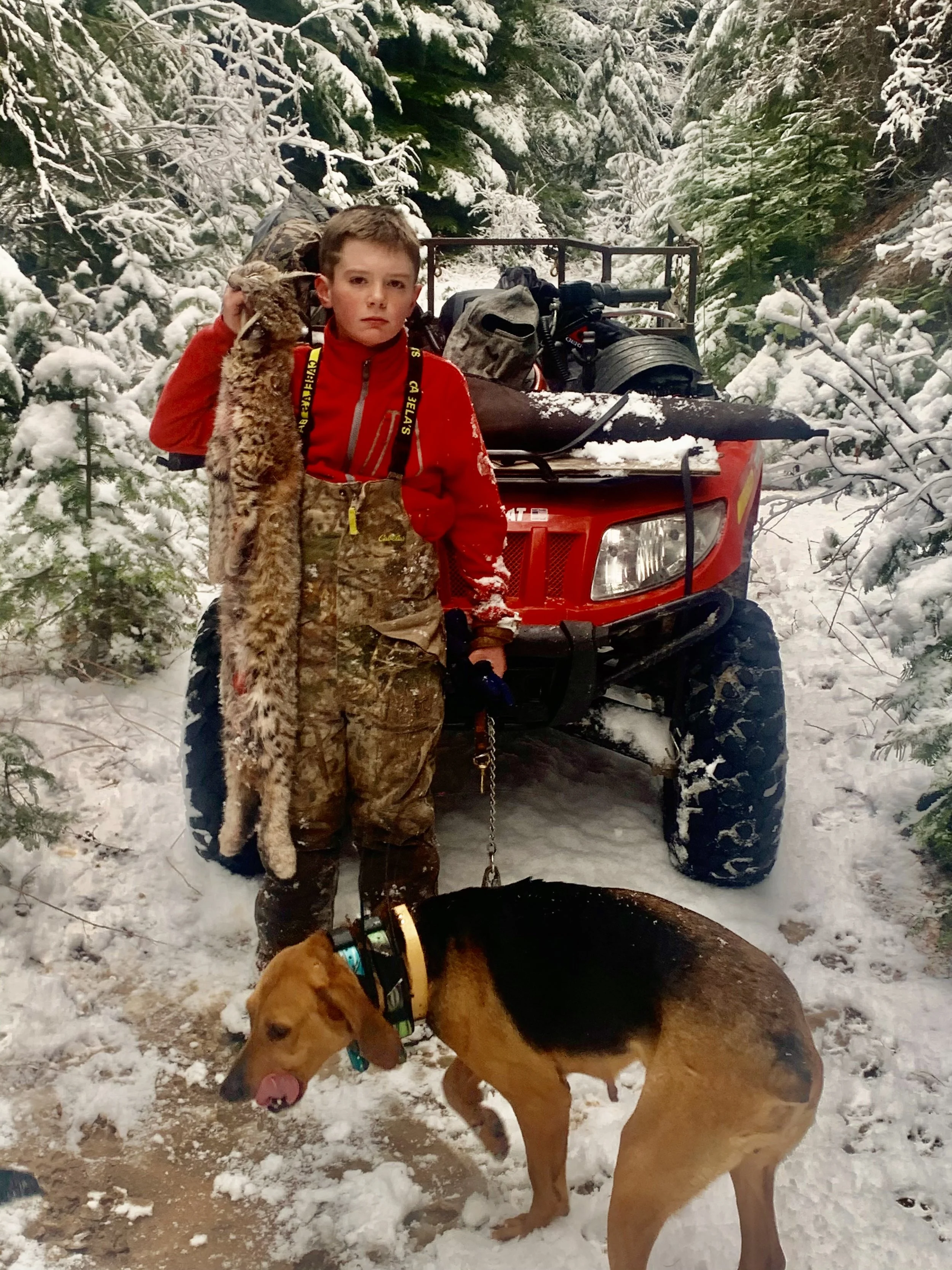 A young boy dressed in camouflage and red outdoor gear holding a bobcat on a leash, standing in snowy forest next to a red all-terrain vehicle with gear. A dog with a collar is sniffing the snow nearby.