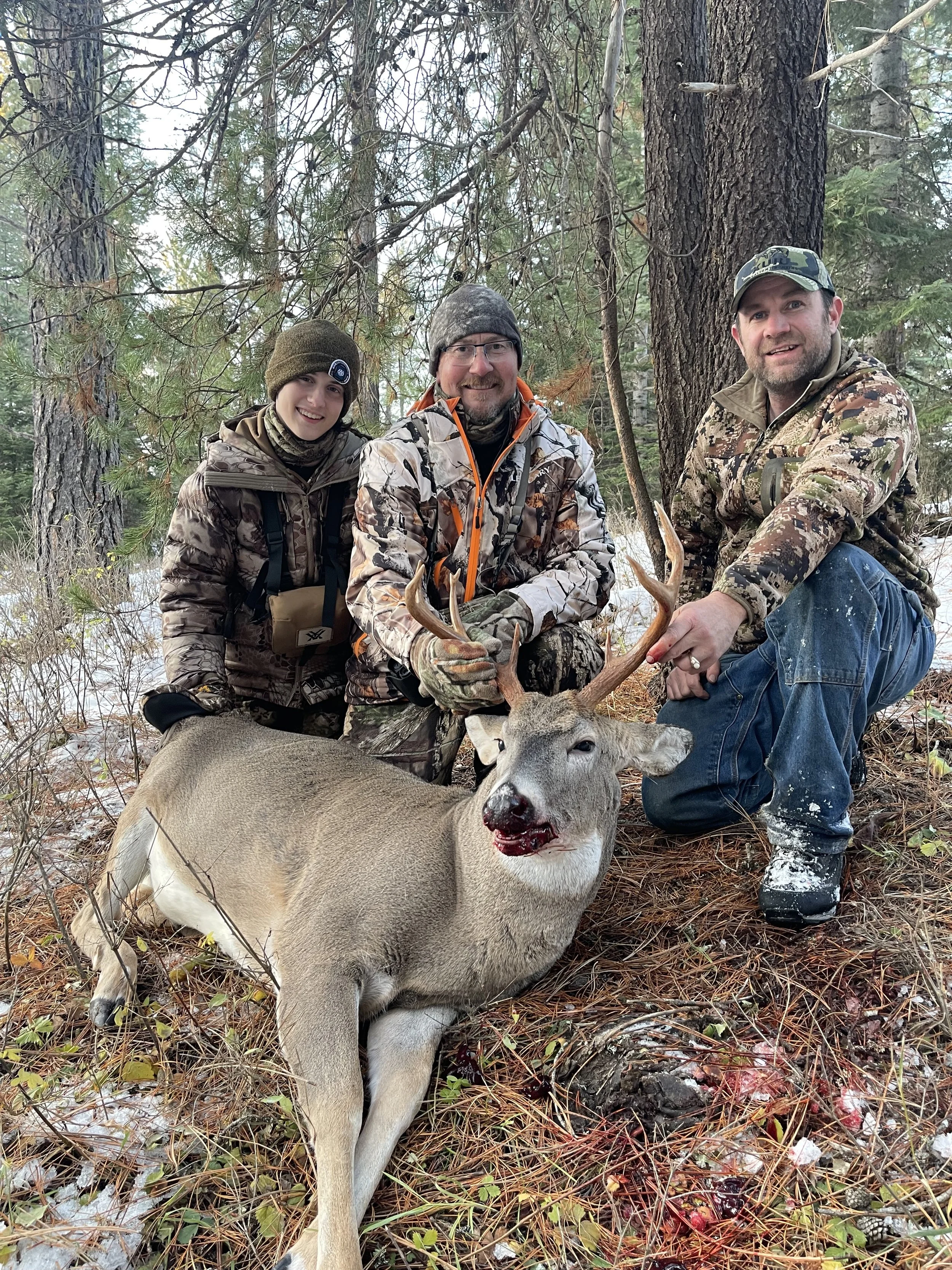 Three hunters in camouflage clothing kneel behind a deer with antlers in a forest during winter, smiling at the camera.